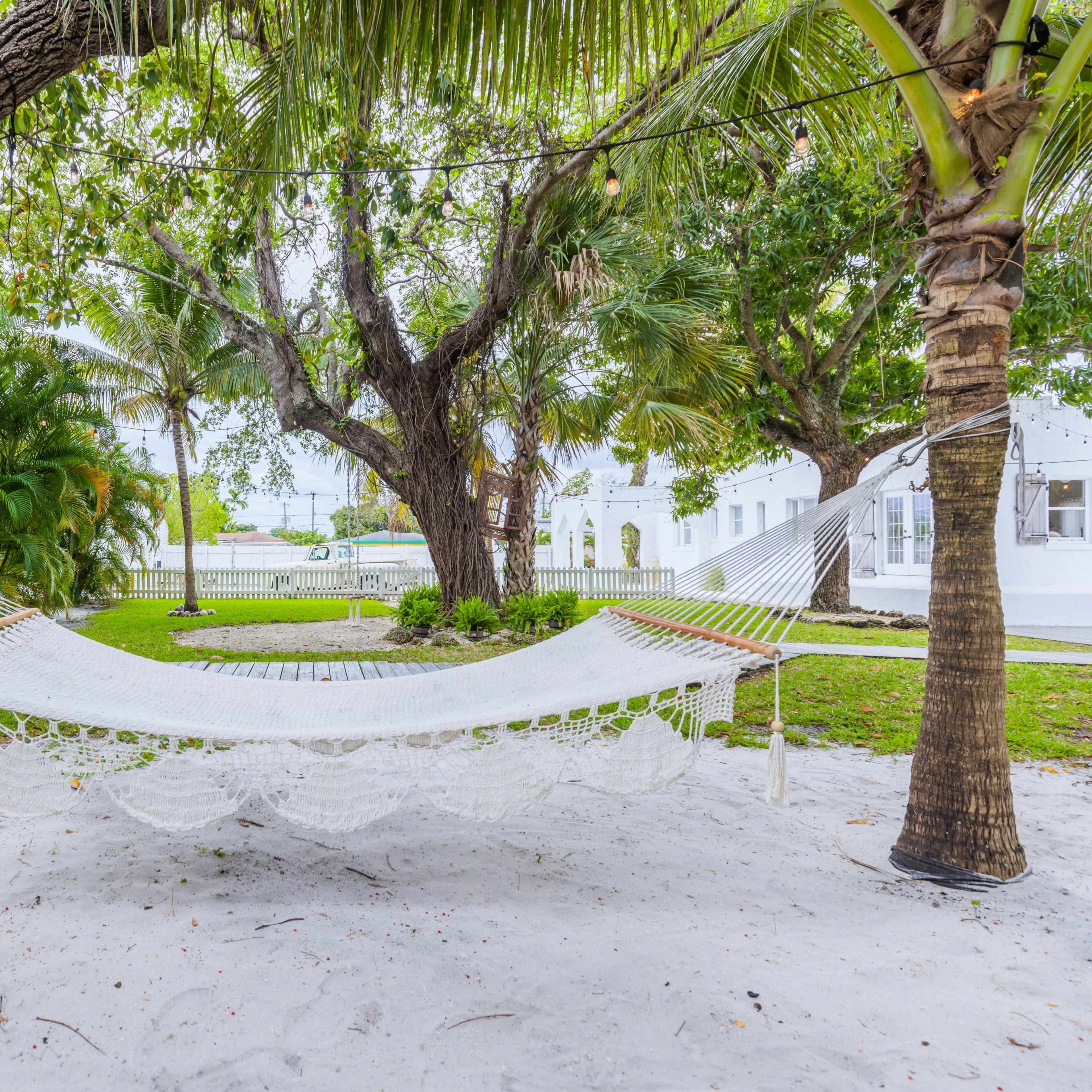 A hammock is strung between two palm trees in a sandy garden surrounded by lush greenery.