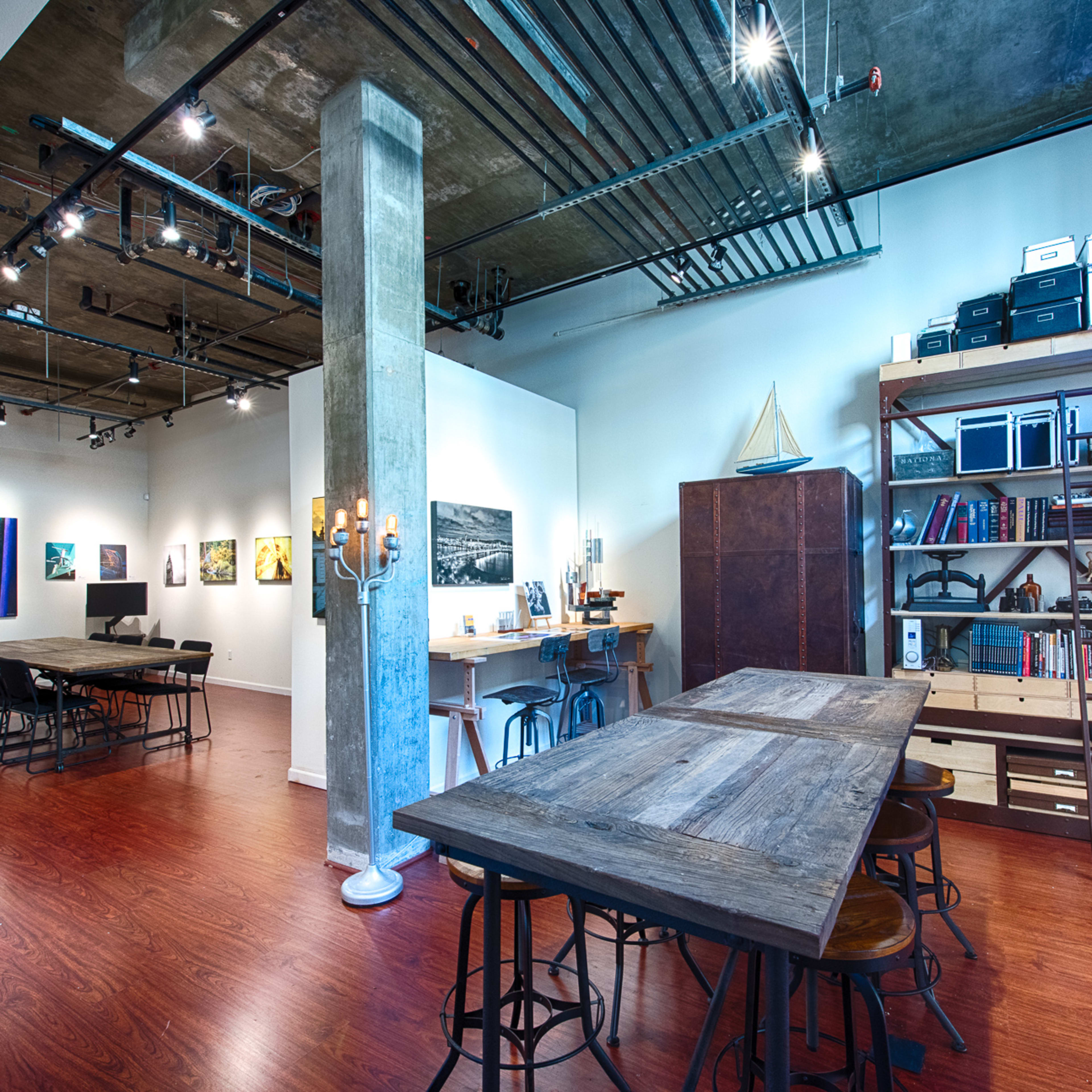 An open studio space featuring a wooden table with stools, shelving units filled with books, artwork displayed on the walls, and a concrete pillar in the center.