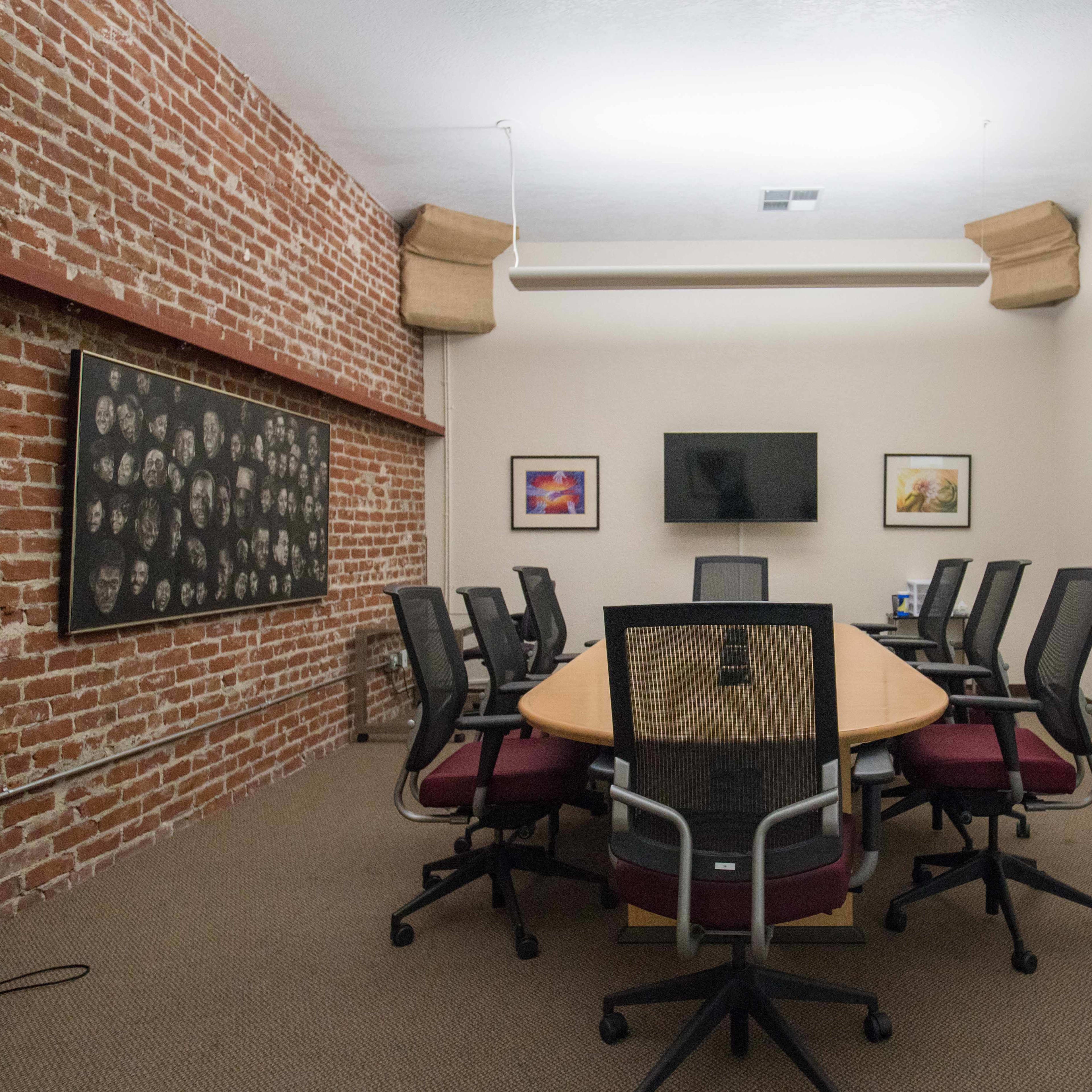 A conference room features a round table surrounded by ergonomic chairs, a television on the wall, a whiteboard, and exposed brick walls.