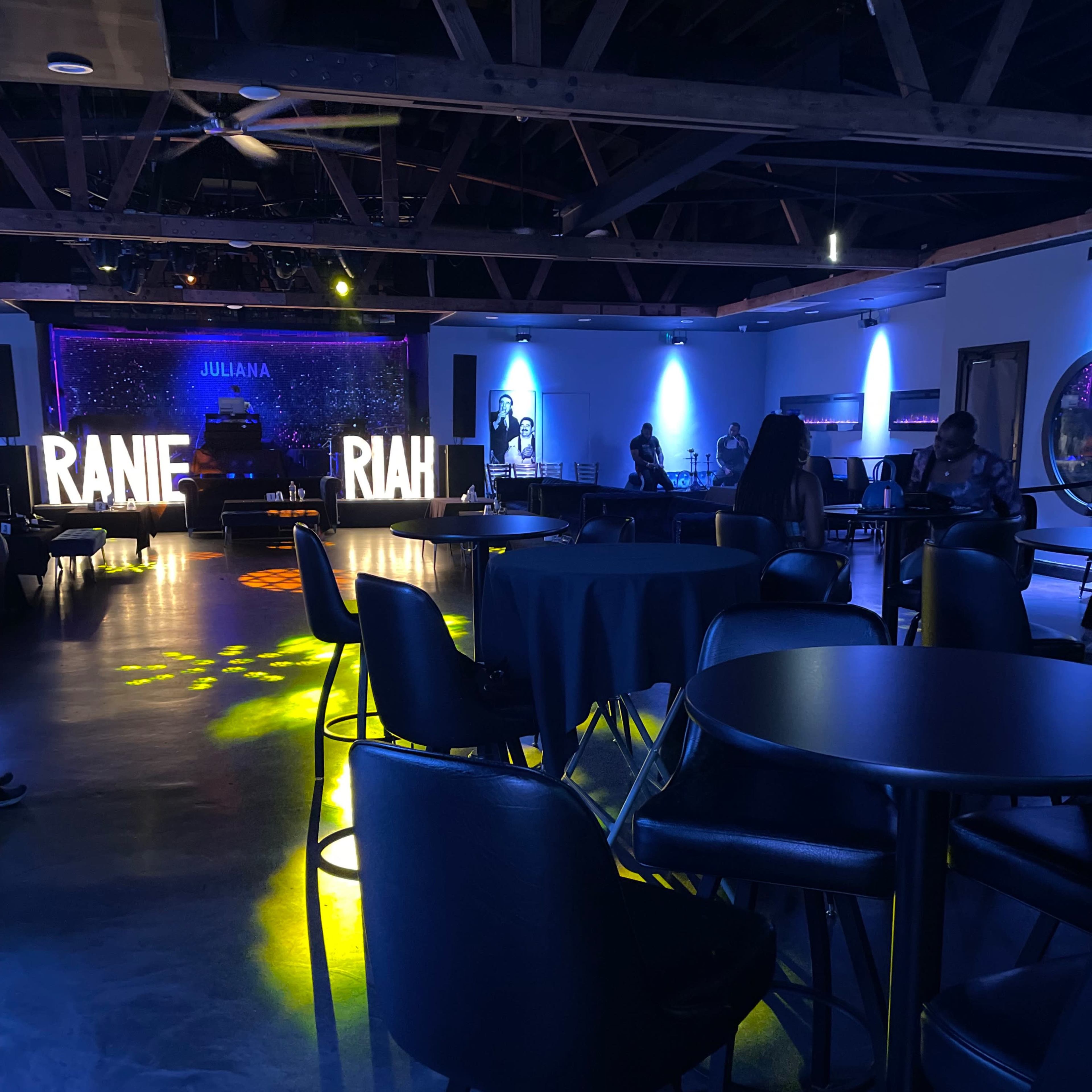 A dimly lit lounge or event space with tables and chairs arranged around a stage featuring illuminated letters spelling "RANIE" and "RIAH."