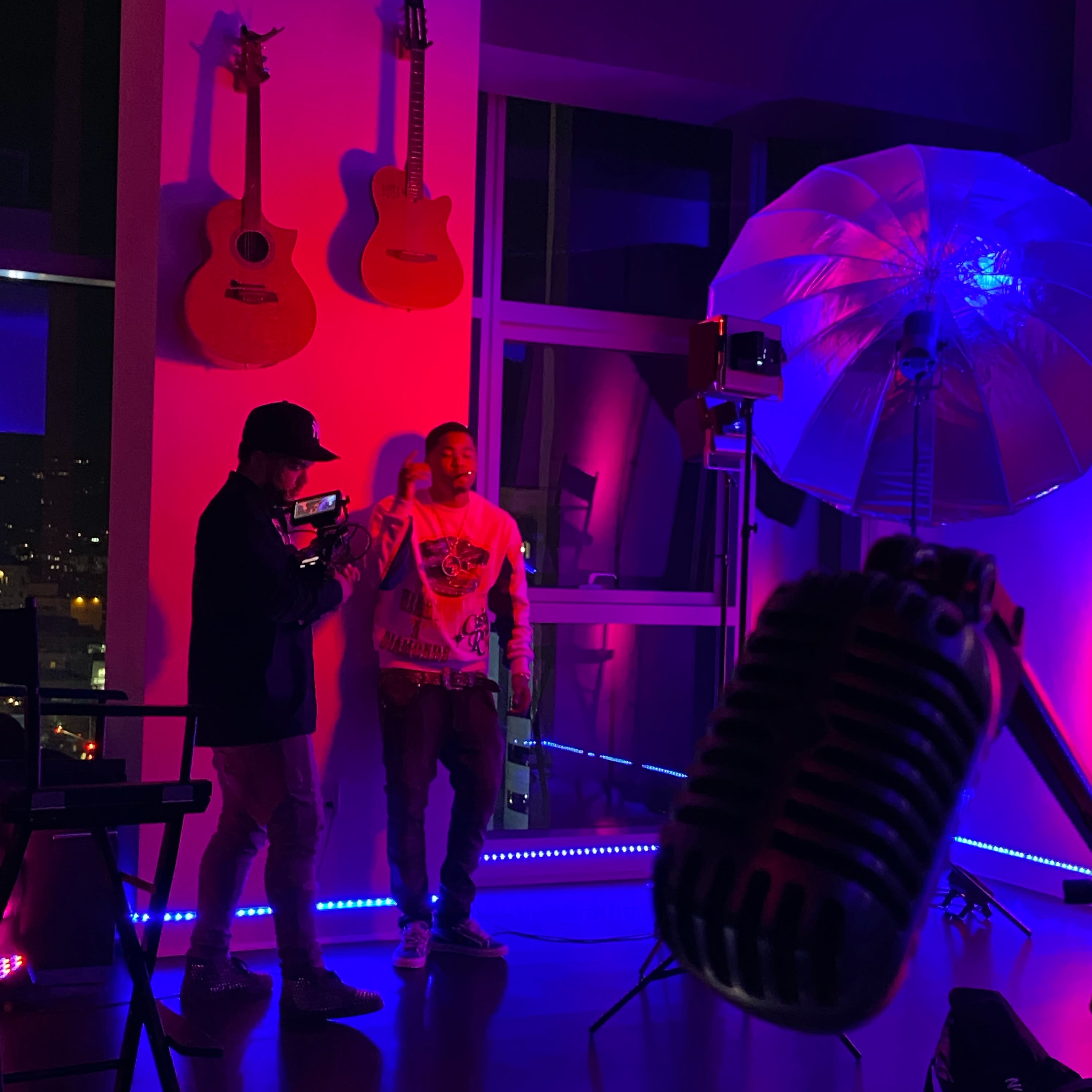 Two men stand in a brightly lit room with guitars mounted on the wall, while a microphone and large umbrella light are positioned nearby.