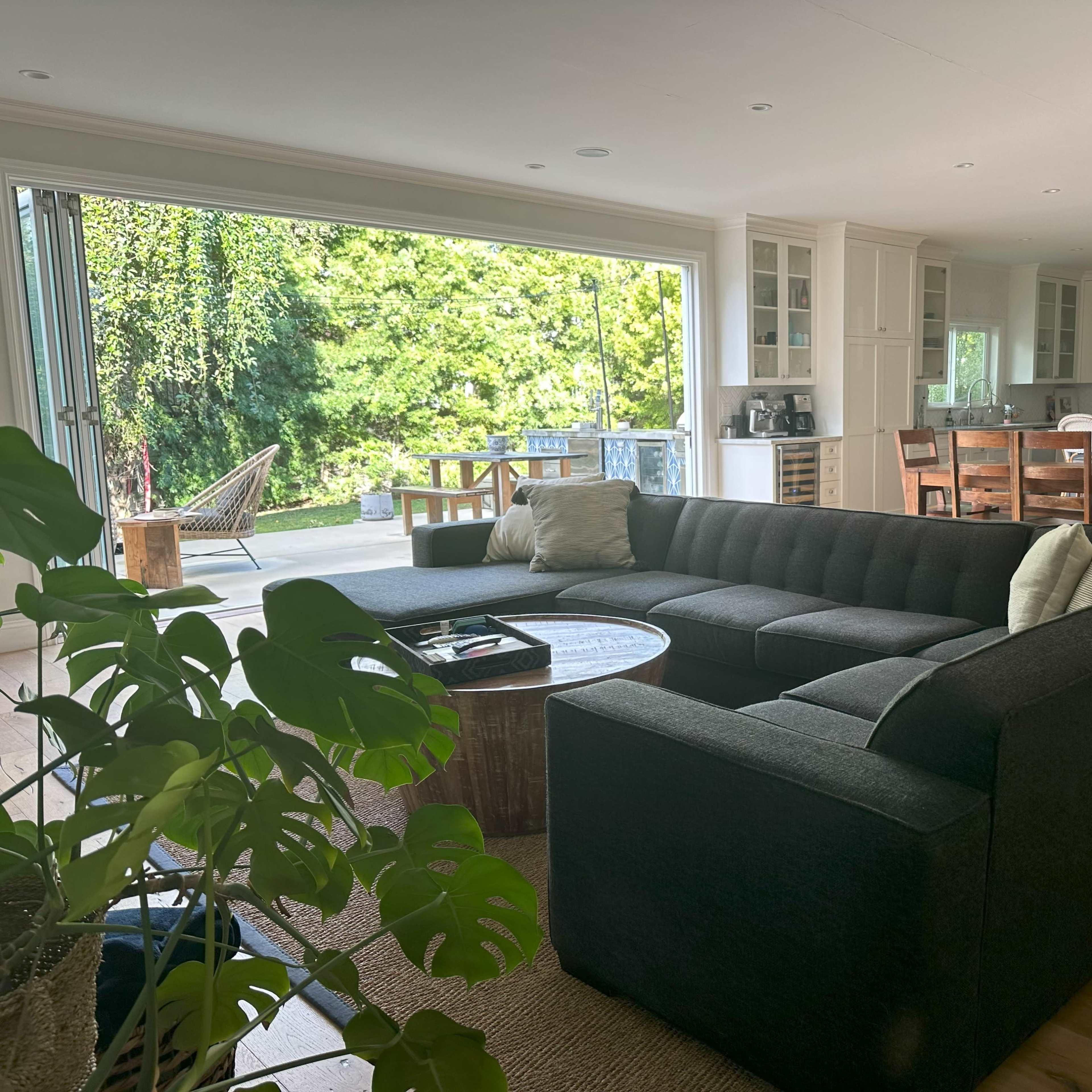 A modern living room with a dark sectional sofa, a round coffee table, and large sliding glass doors that open to an outdoor deck and garden view.