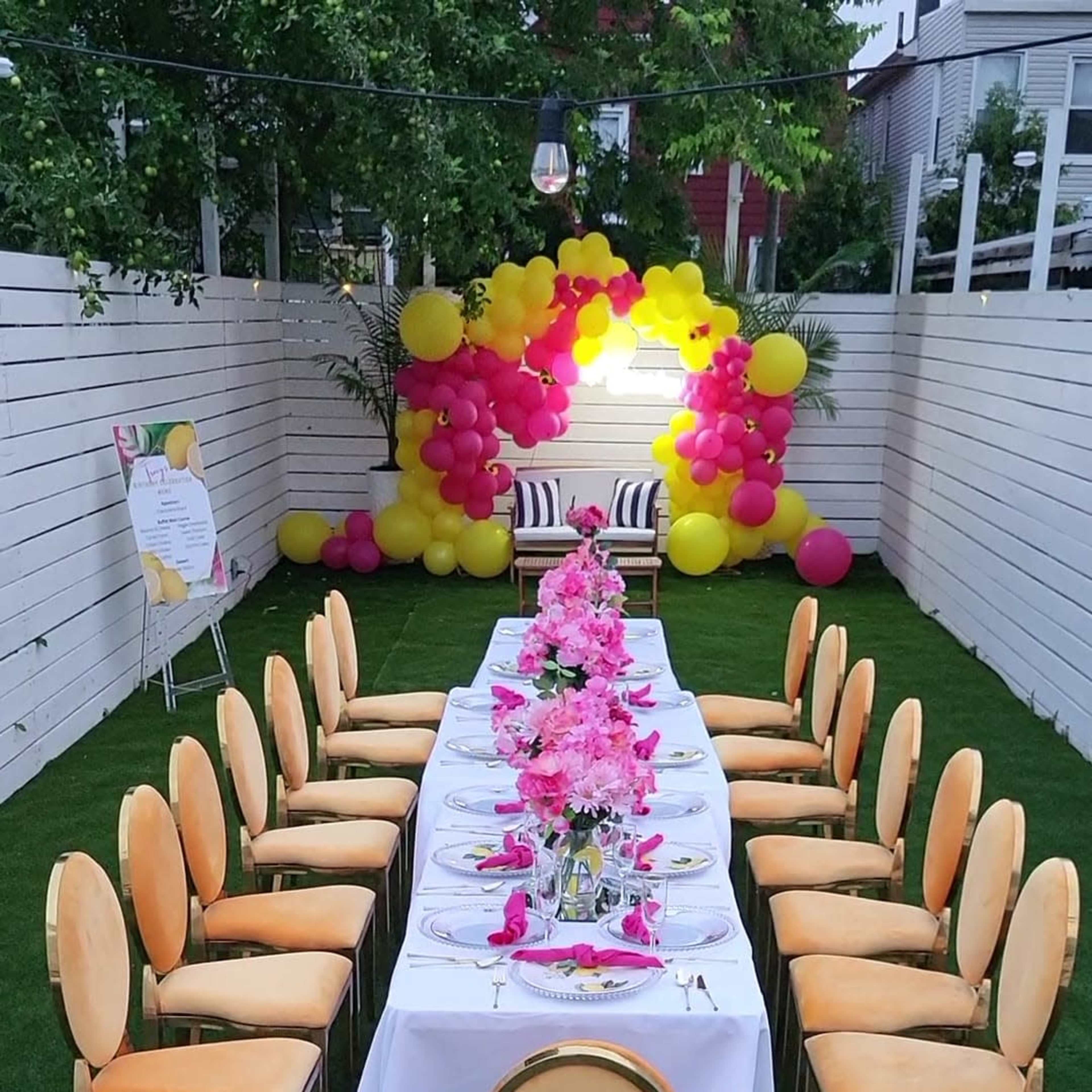 A long dining table is set up outdoors with orange chairs and a floral centerpiece, surrounded by colorful balloons and green grass.