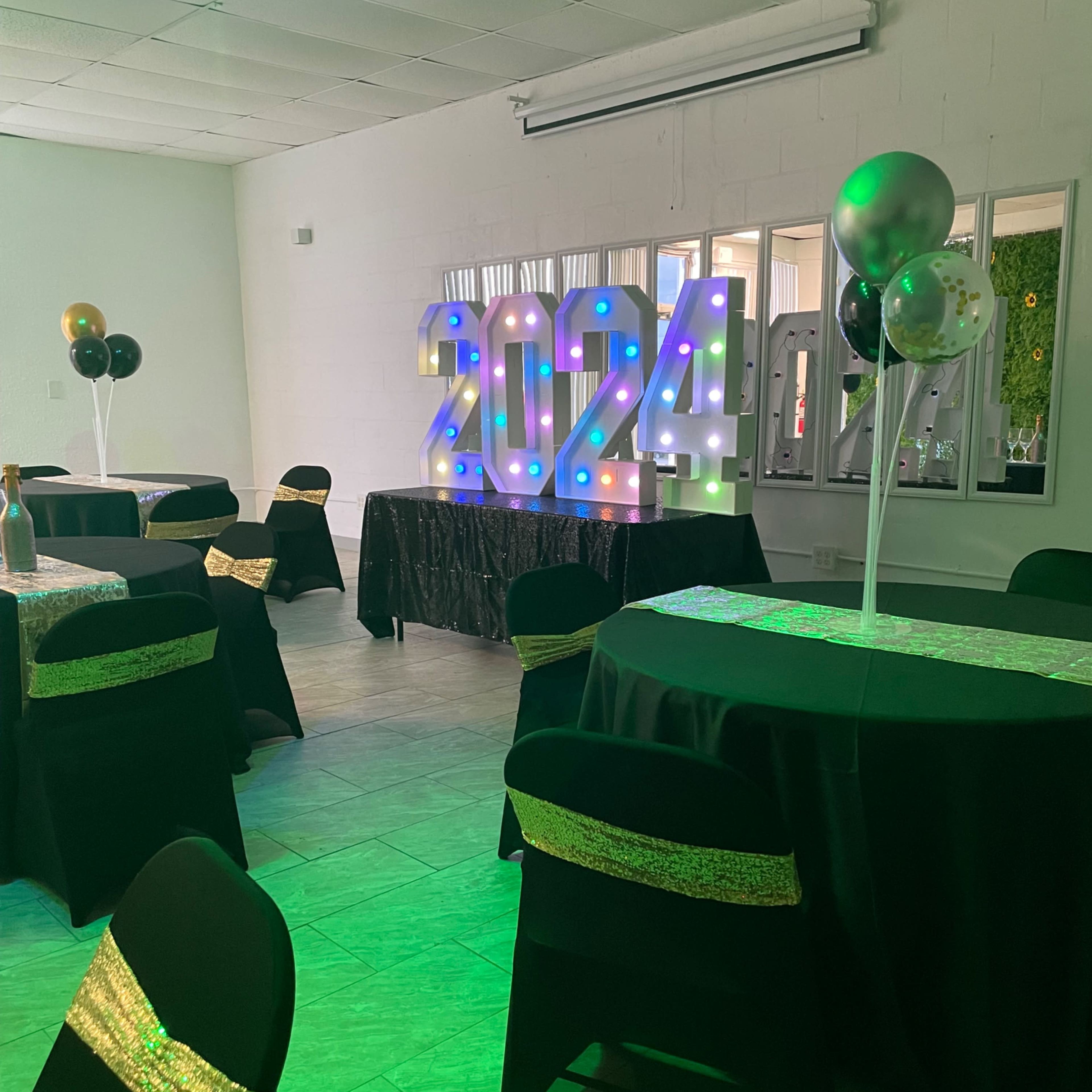 A decorated event space with black tables, chairs covered in black fabric with gold accents, and large illuminated numbers reading "2024" as a backdrop.
