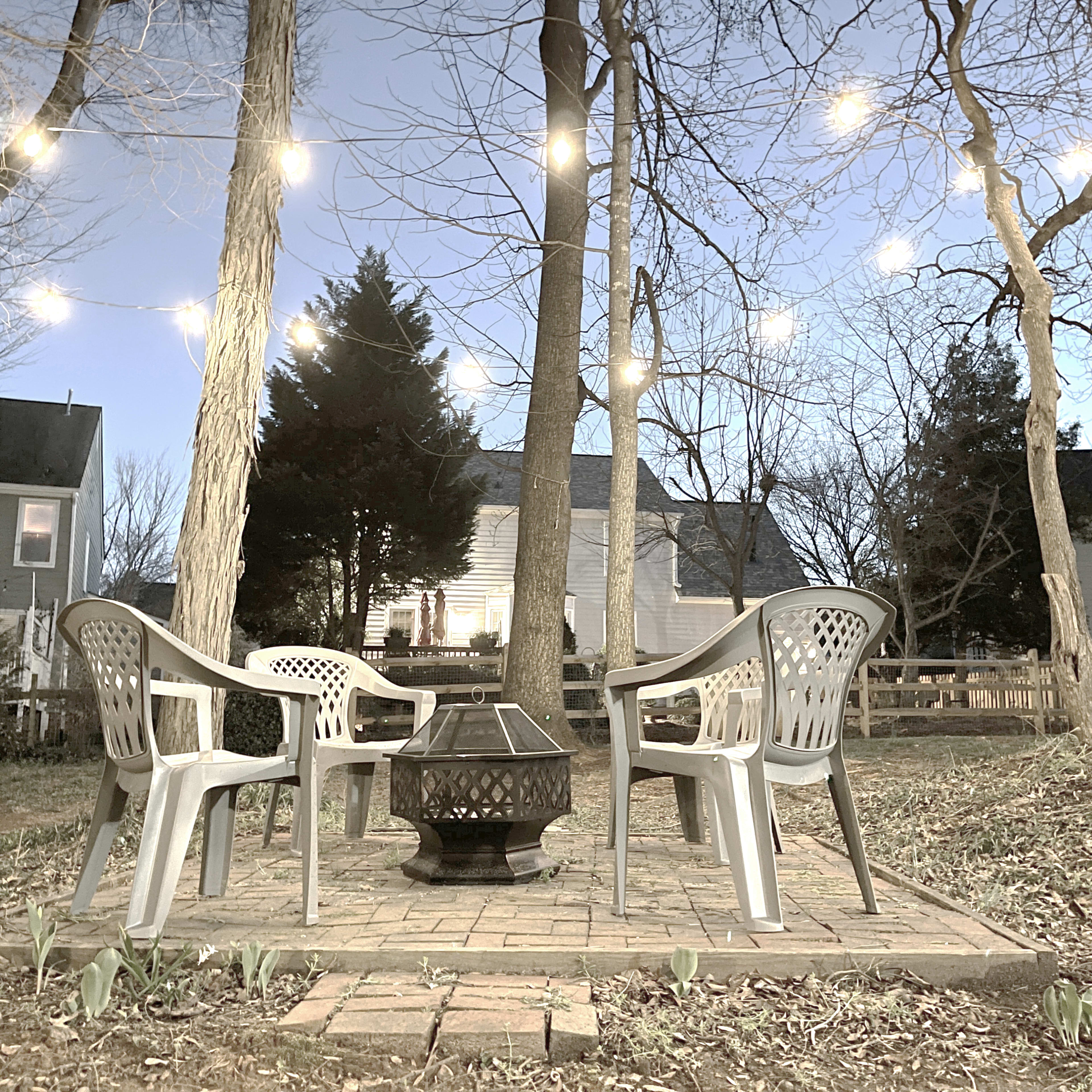 A patio area features four plastic chairs arranged around a fire pit, surrounded by trees draped with string lights.