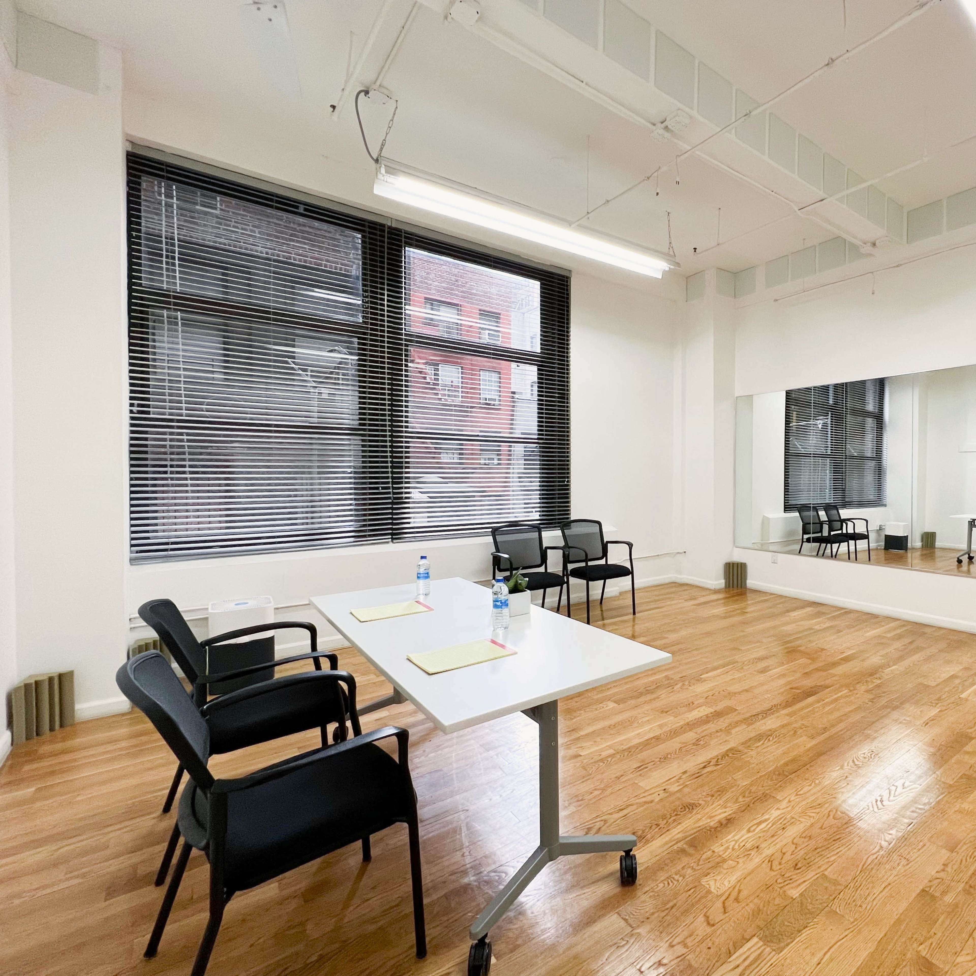 A bright, empty room with hardwood floors, a table with water bottles, and chairs arranged around it, alongside a large mirror and windows with blinds.