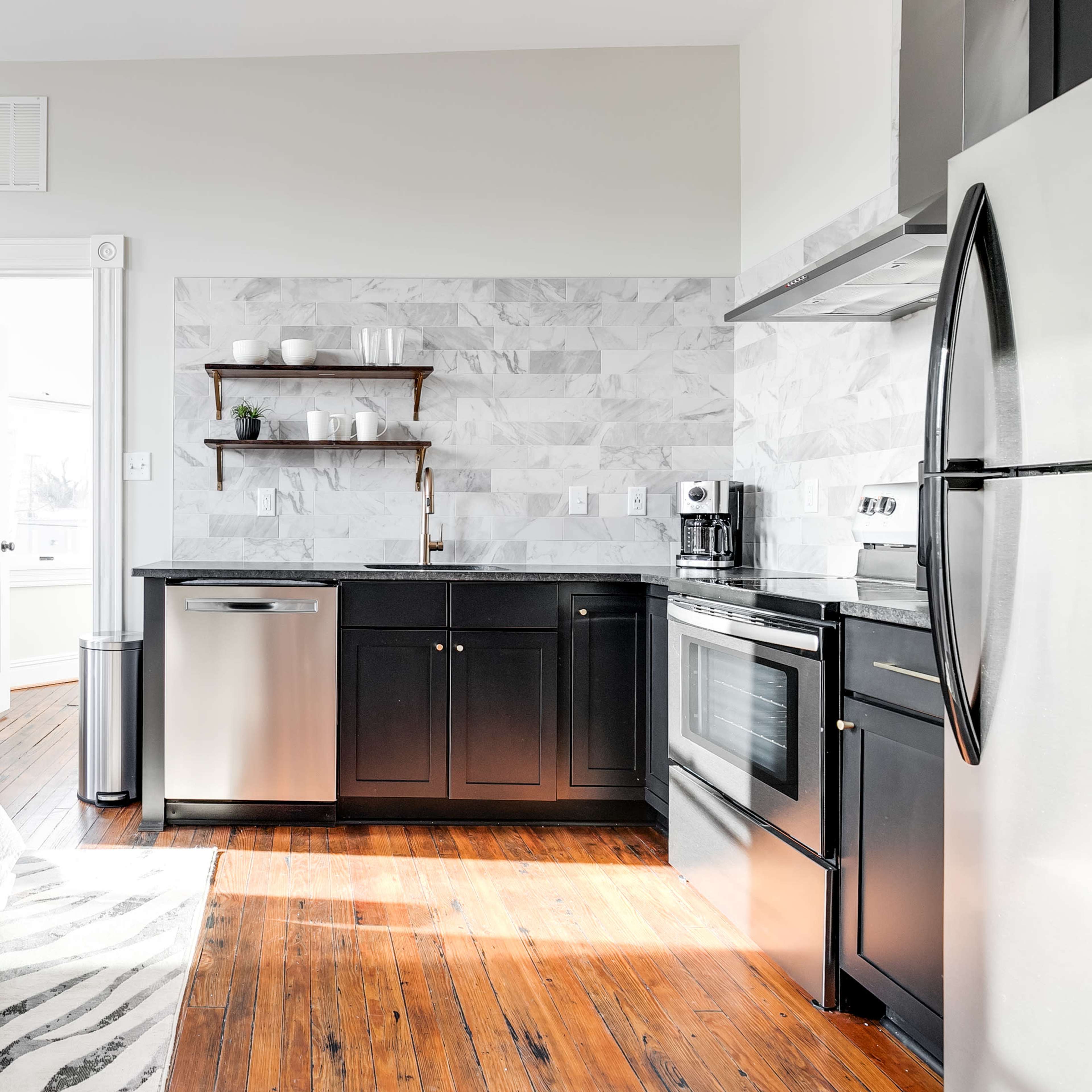 A modern studio apartment featuring a bed in the foreground, a kitchen area with a stainless steel refrigerator and oven, and a door leading to another room.