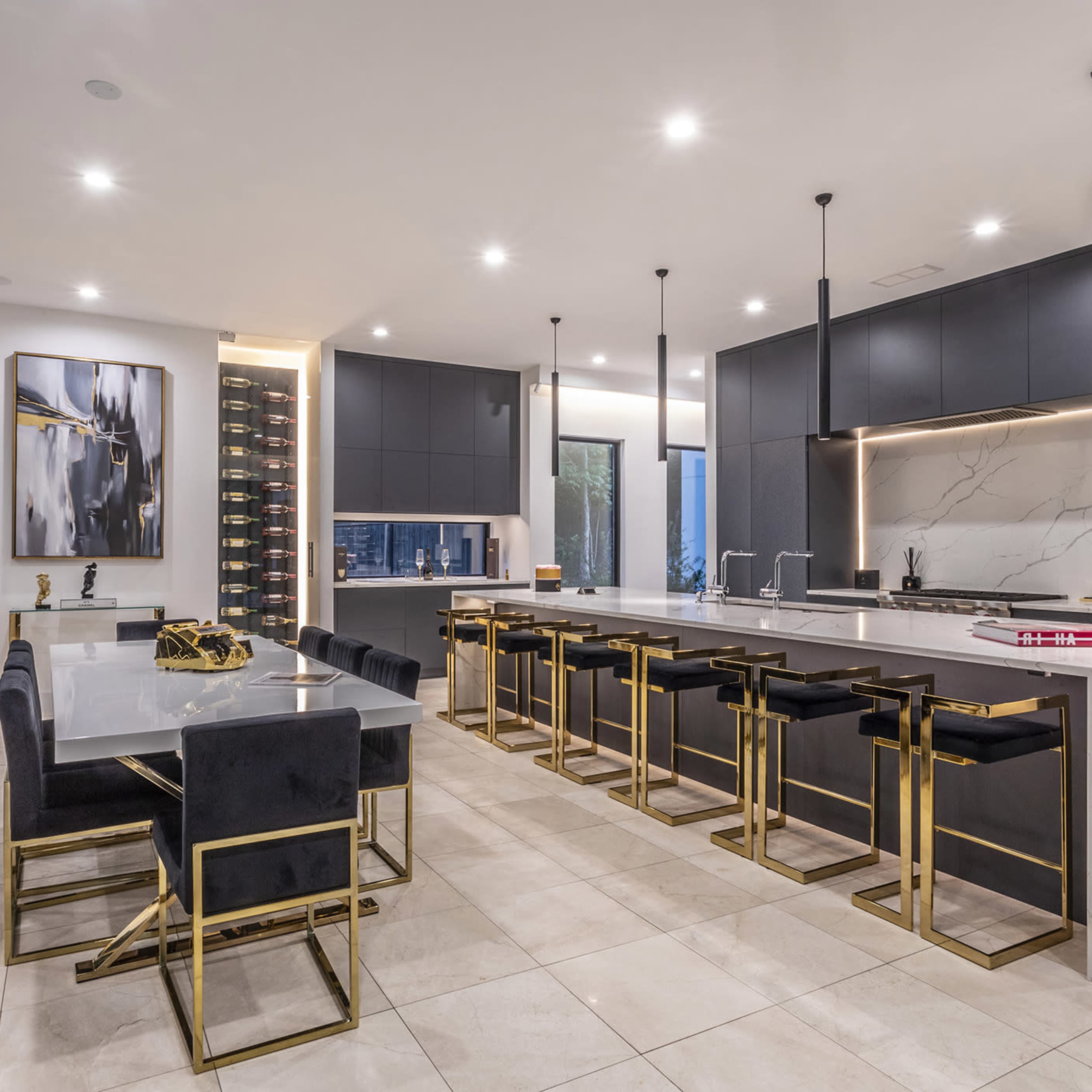 A modern kitchen with dark cabinetry, a large marble island, gold accents, and a dining area featuring a sleek table and chairs.