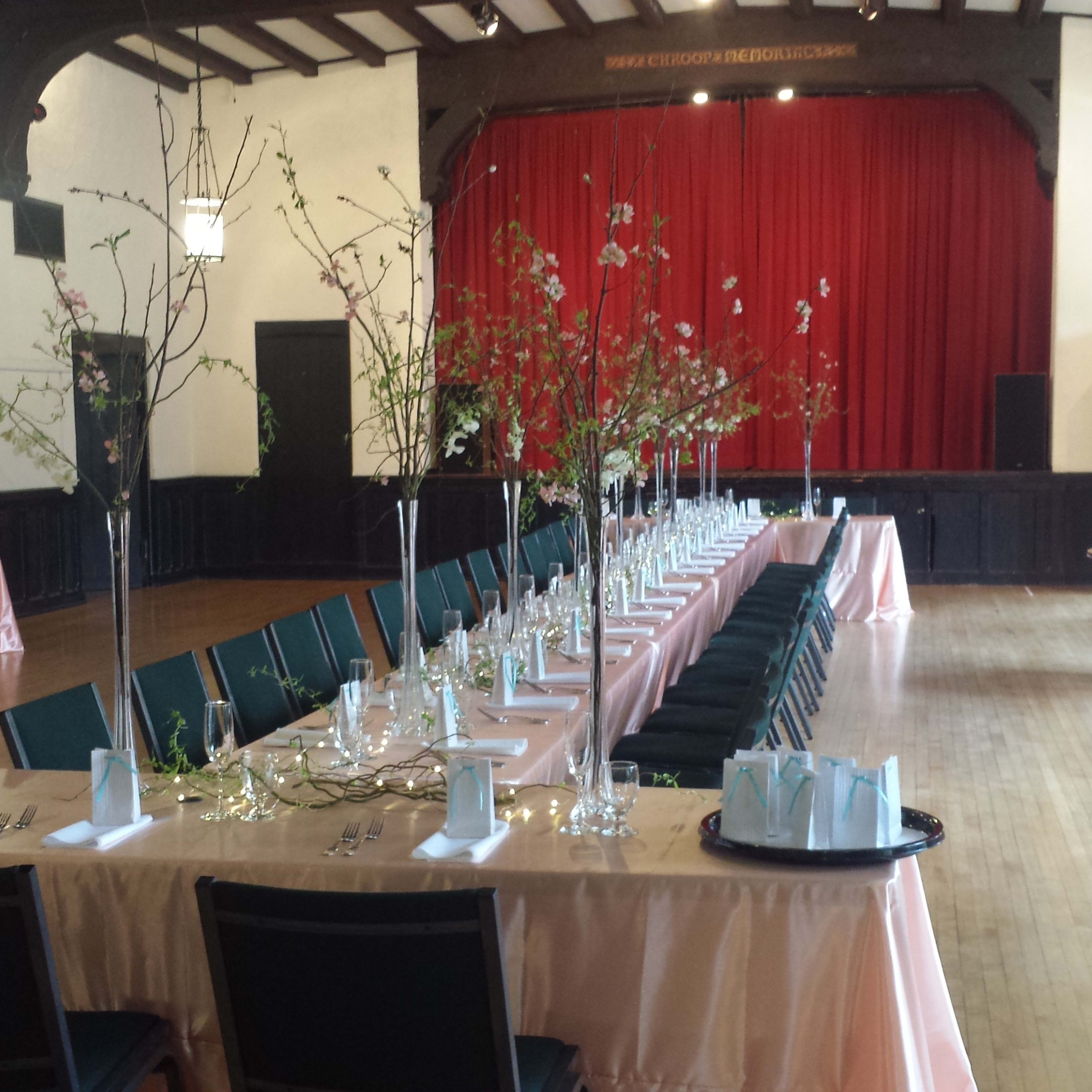 A banquet hall is arranged with long tables dressed in pink tablecloths and decorated with branches and glassware, set against a backdrop of red curtains and wooden beams.