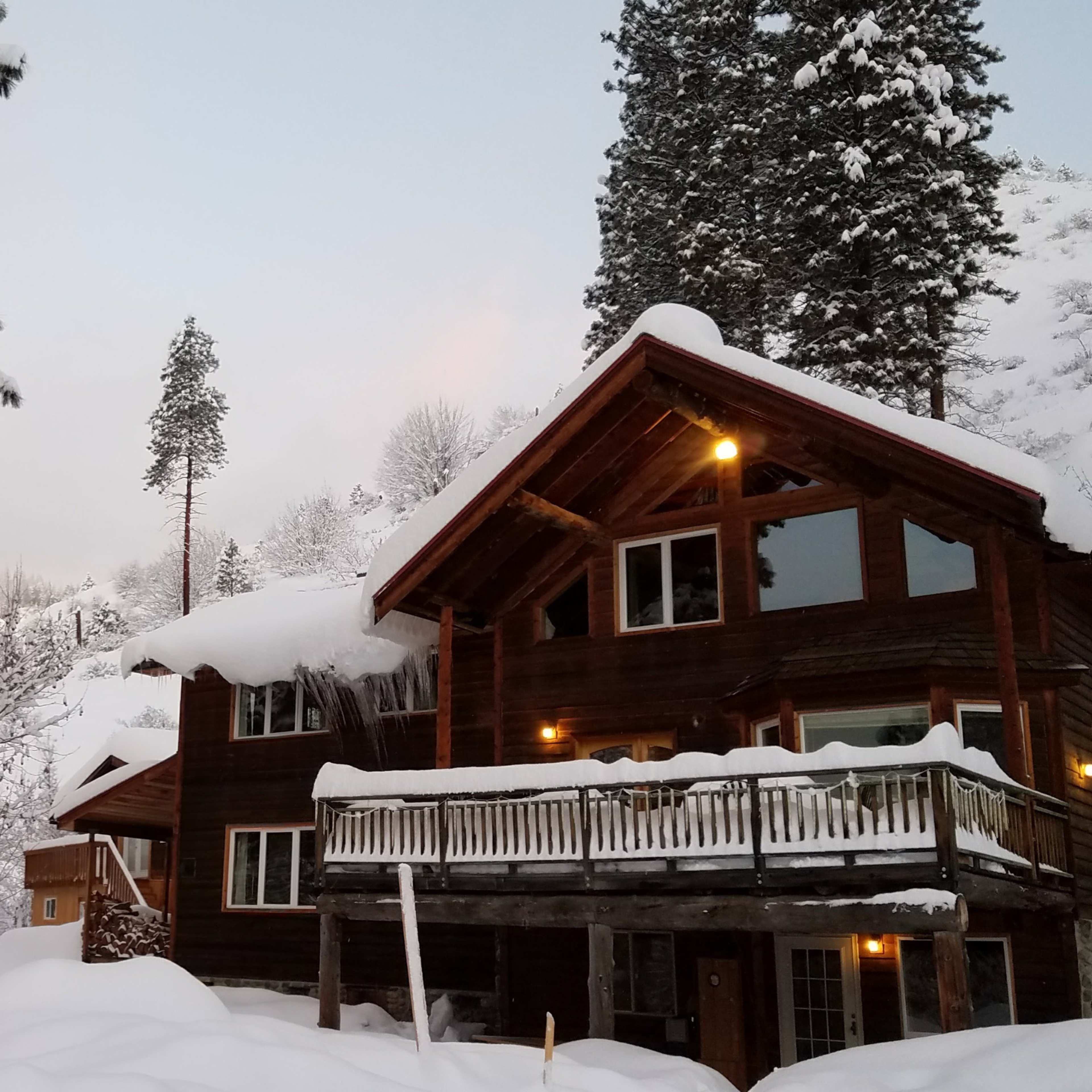 A large wooden cabin is surrounded by snow-covered trees and ground, with warm lights glowing from its windows.