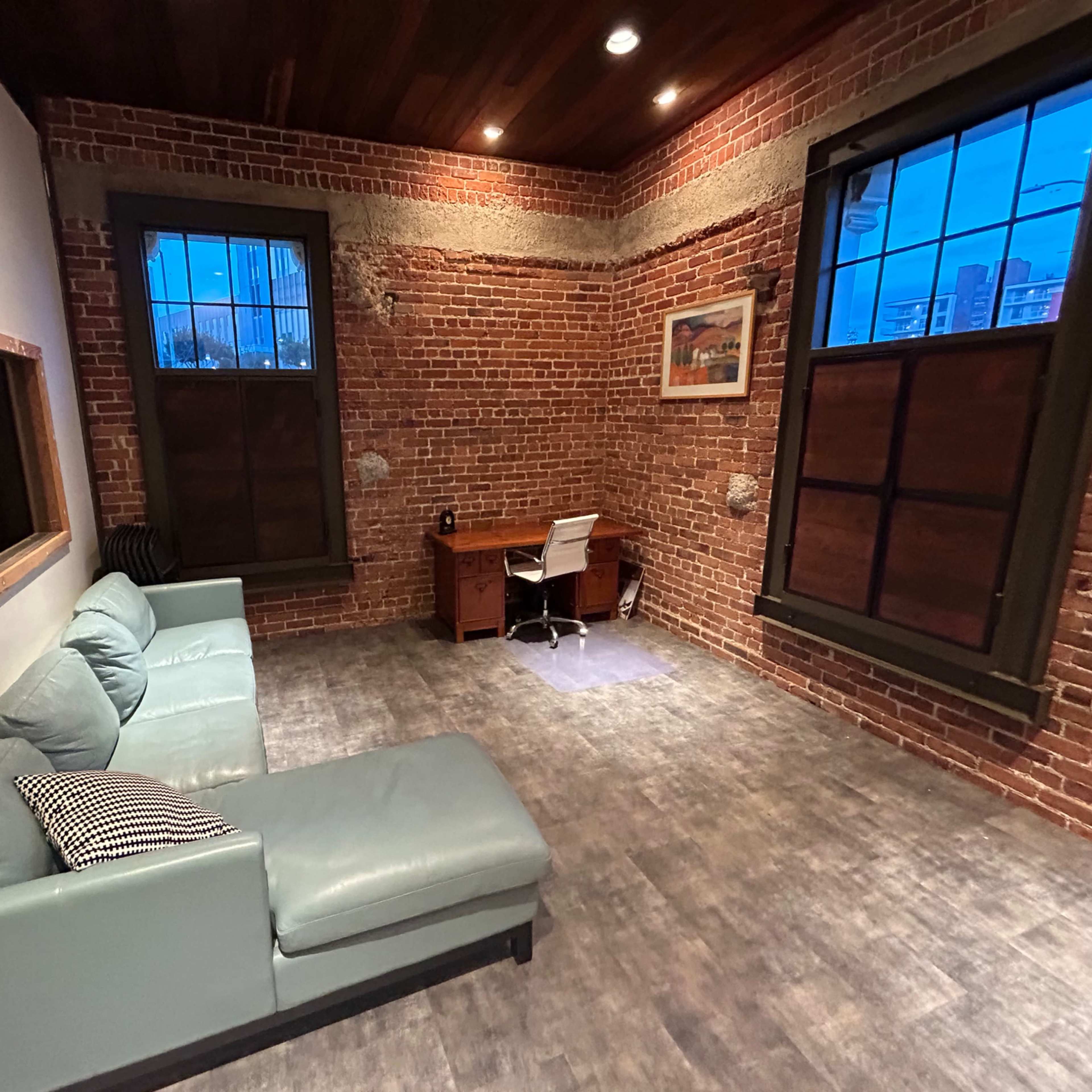 The room features exposed brick walls, a green sectional sofa, a wooden desk, and large windows allowing natural light.