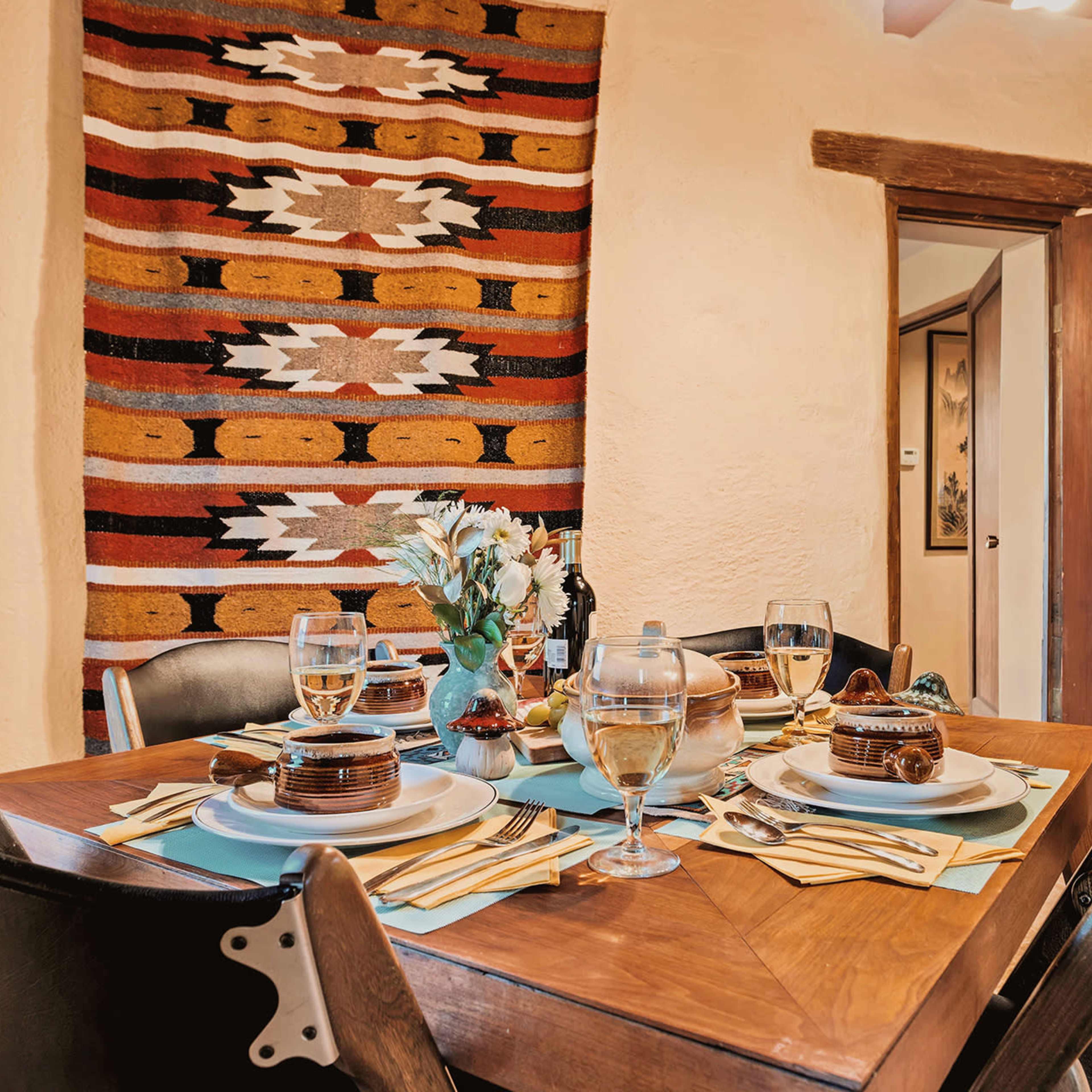 A dining table is set with glasses, plates, and utensils, surrounded by chairs, with a decorative woven tapestry on the wall.