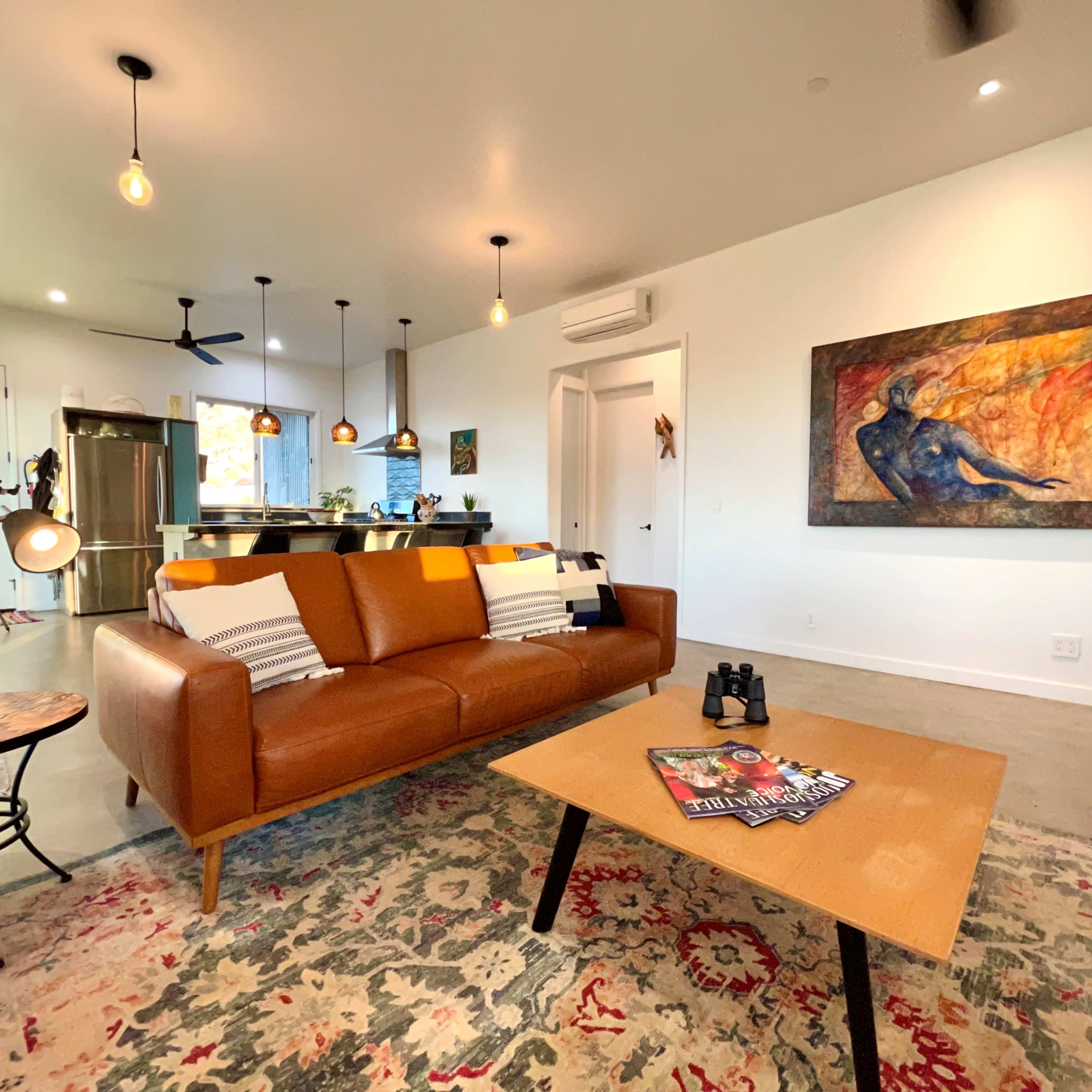 The living area features a brown leather couch, a coffee table with magazines, and an abstract painting on the wall, accompanied by modern light fixtures.
