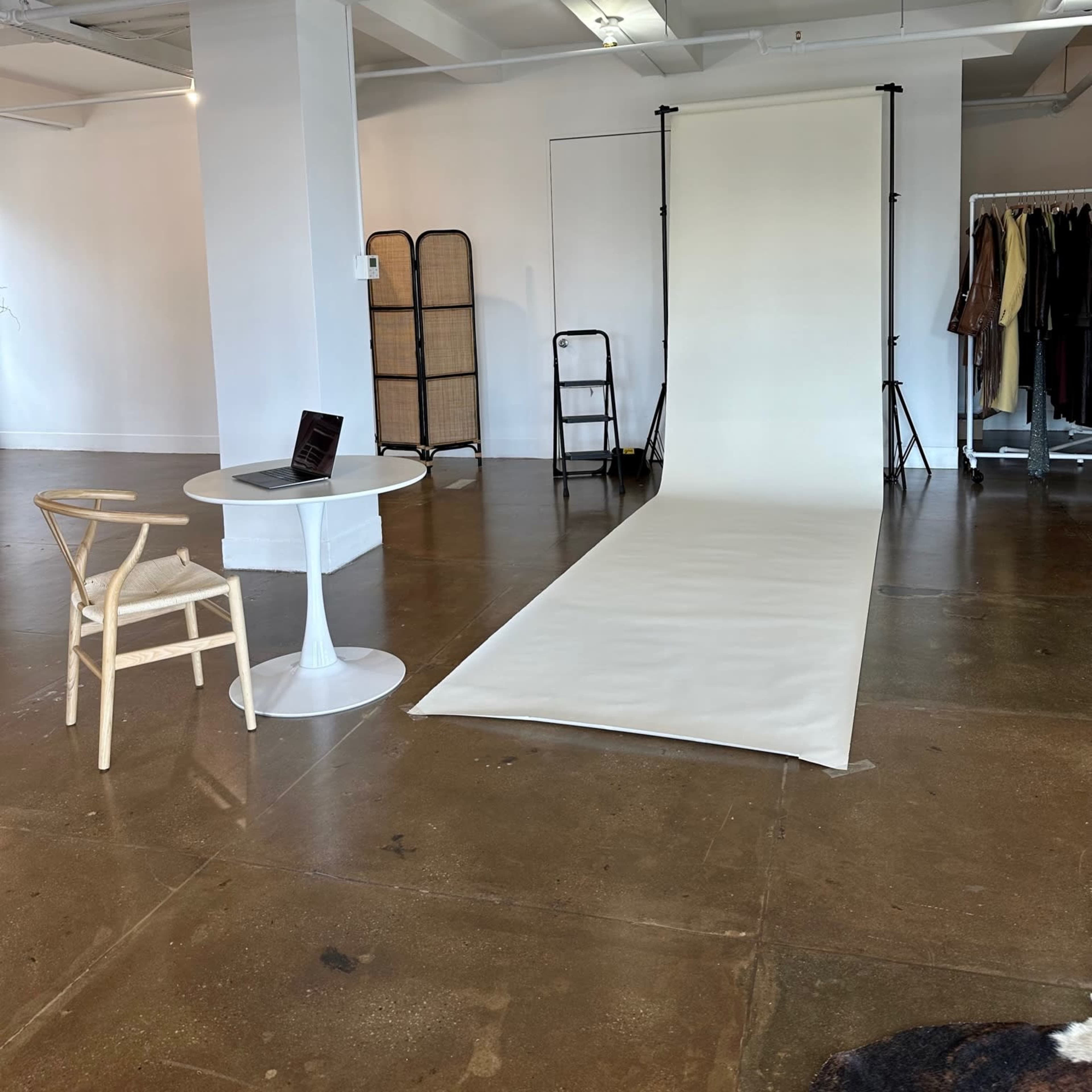 A simple studio space featuring a white backdrop setup, a white table with a laptop, and a clothing rack in the background.