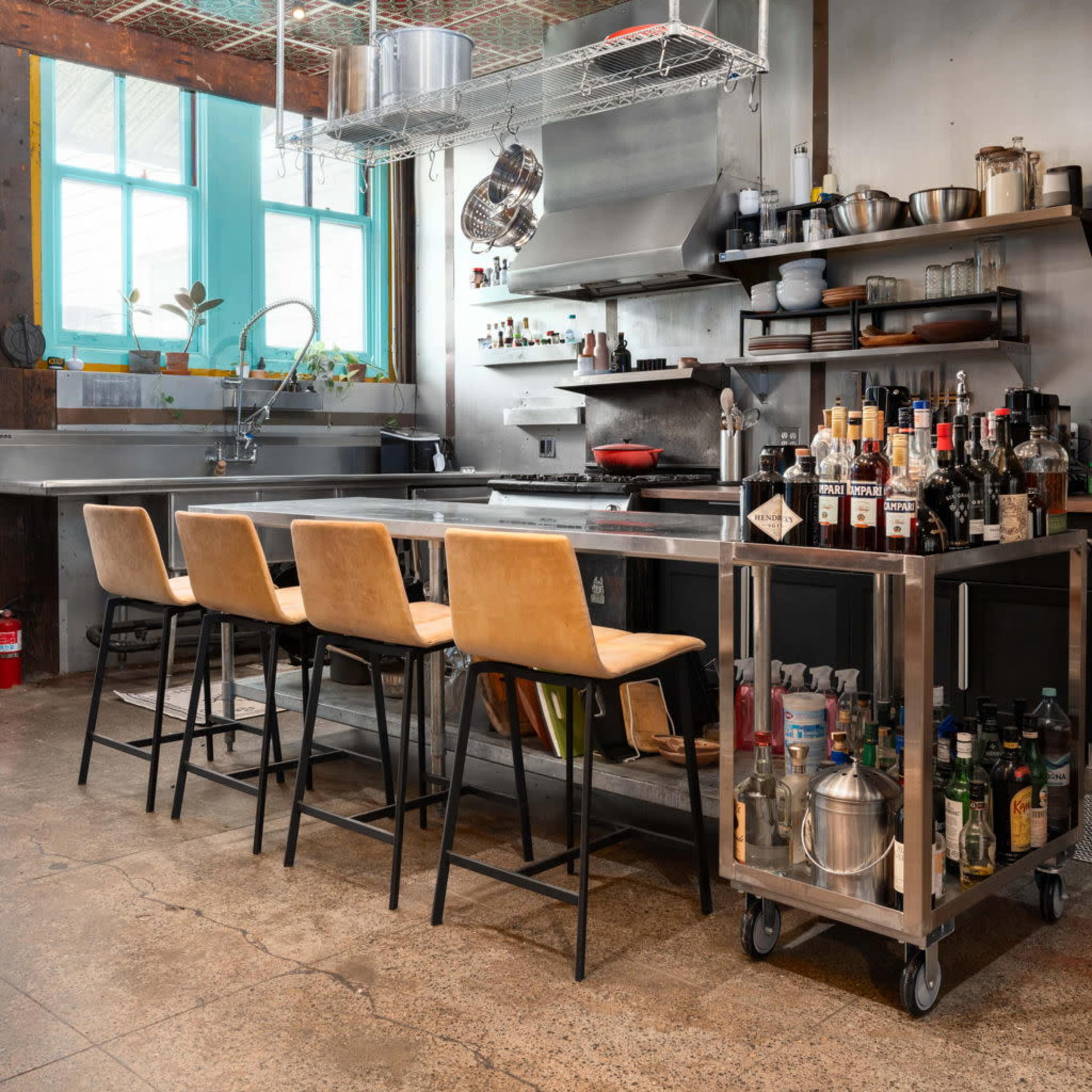 A modern kitchen with a central island, four bar stools, stainless steel appliances, and shelves filled with various kitchen tools and bottles.