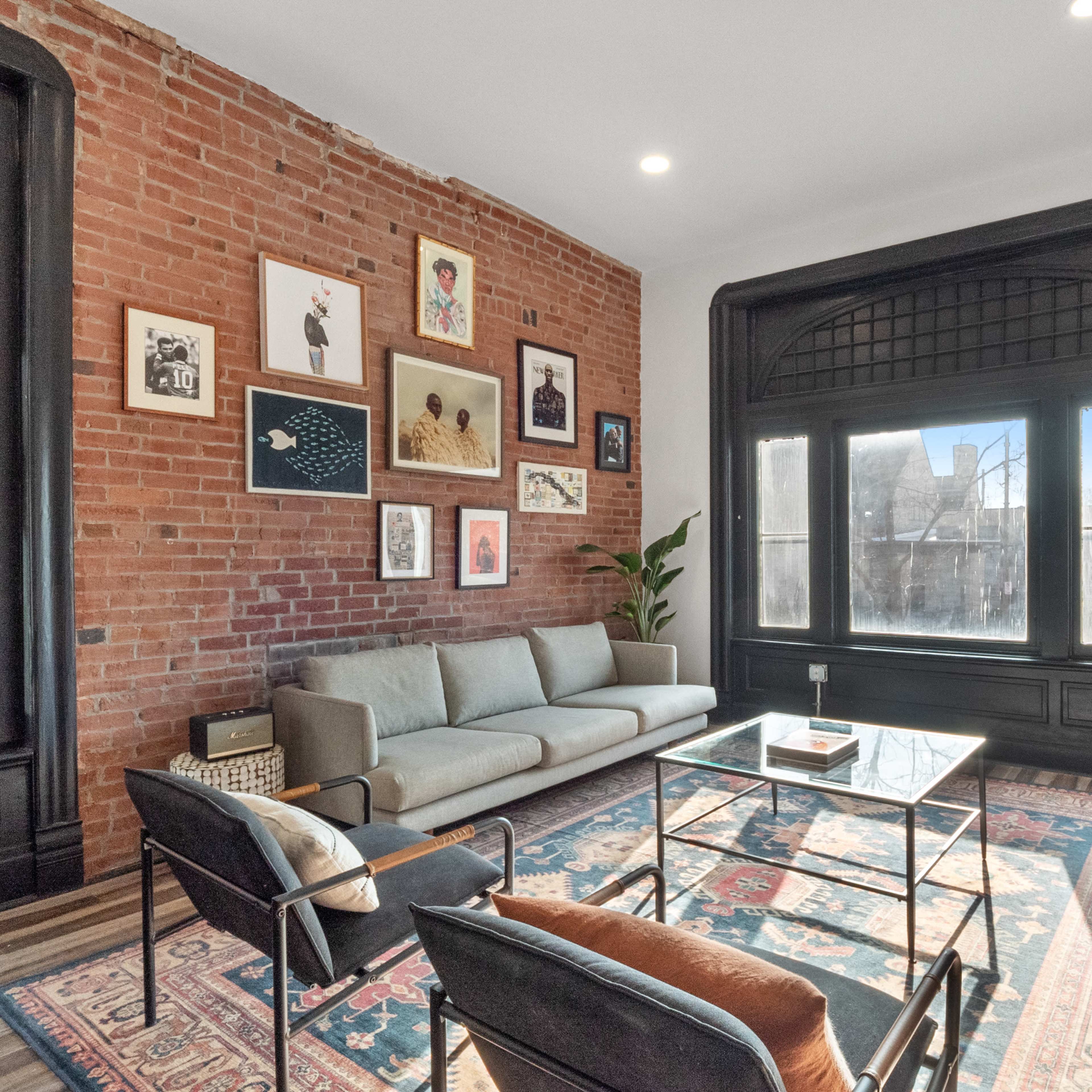 A modern living room with exposed brick walls, a green sofa, a blue couch, a glass coffee table, and a large window alongside framed artwork.