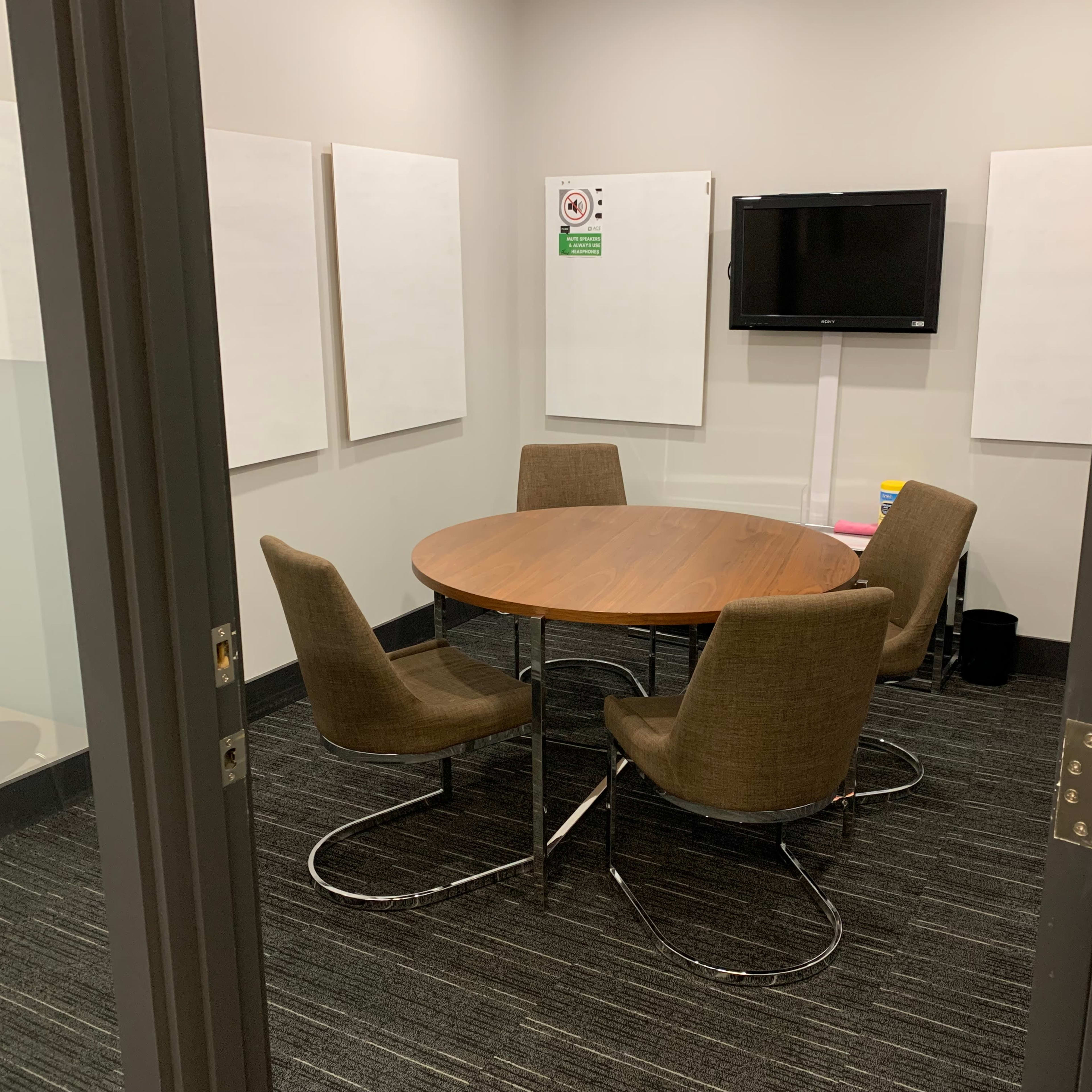 A small meeting room with a round table and four chairs, a television mounted on the wall, and several blank whiteboards.
