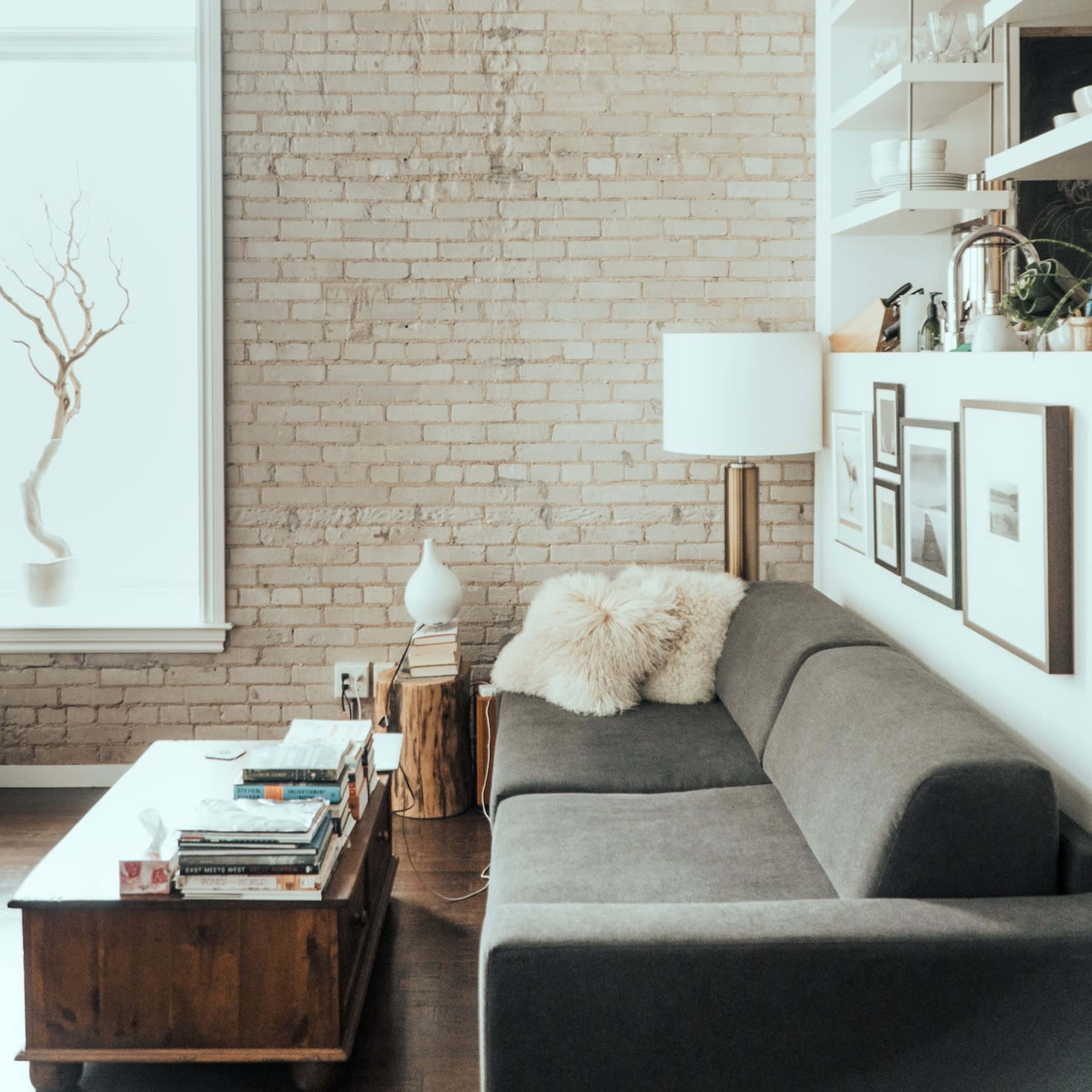 A cozy living room with a gray sofa, a wooden coffee table stacked with books, and a large window with natural light illuminating a brick wall.
