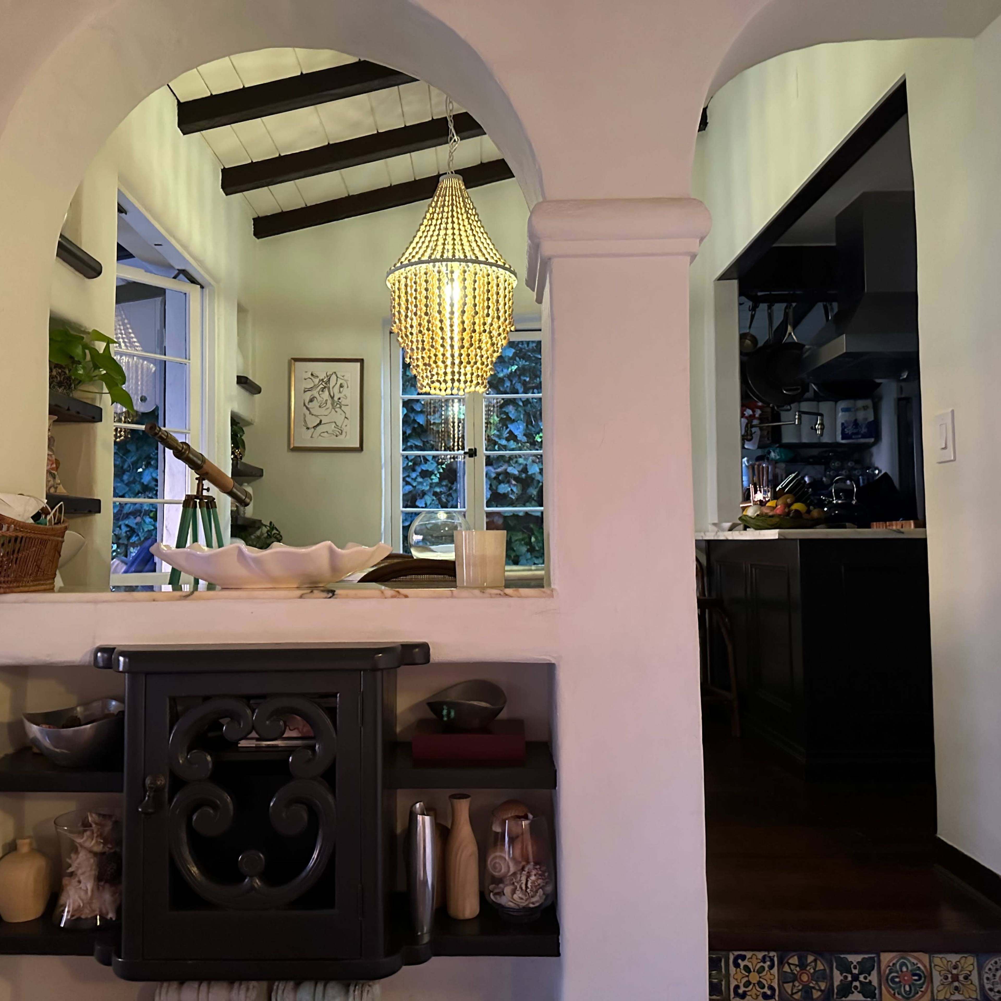 A view of a room with an arched doorway leading to a kitchen, featuring a decorative chandelier and a shelf filled with various items.