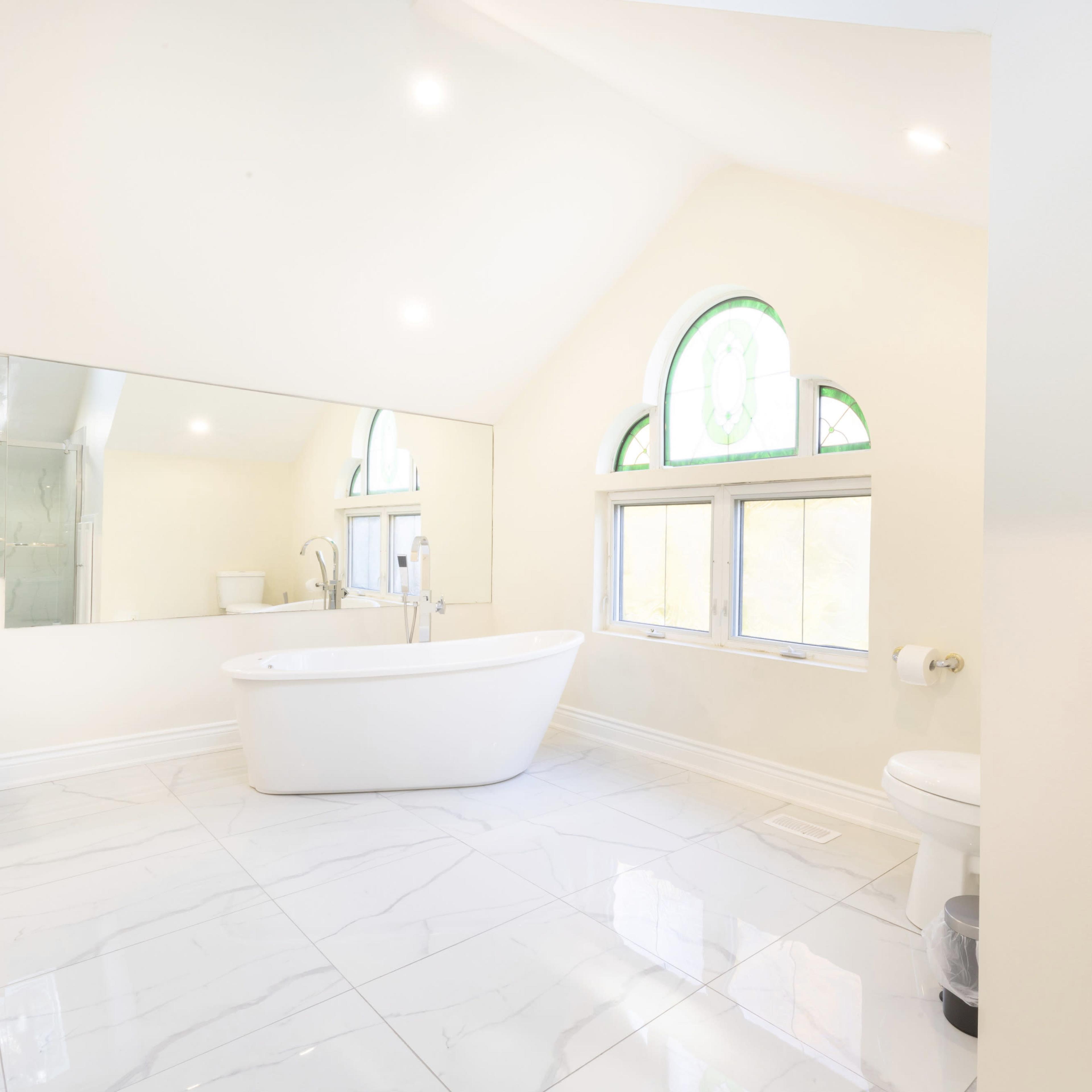 A spacious, modern bathroom with a freestanding bathtub, large mirror, and a window featuring an arched design.