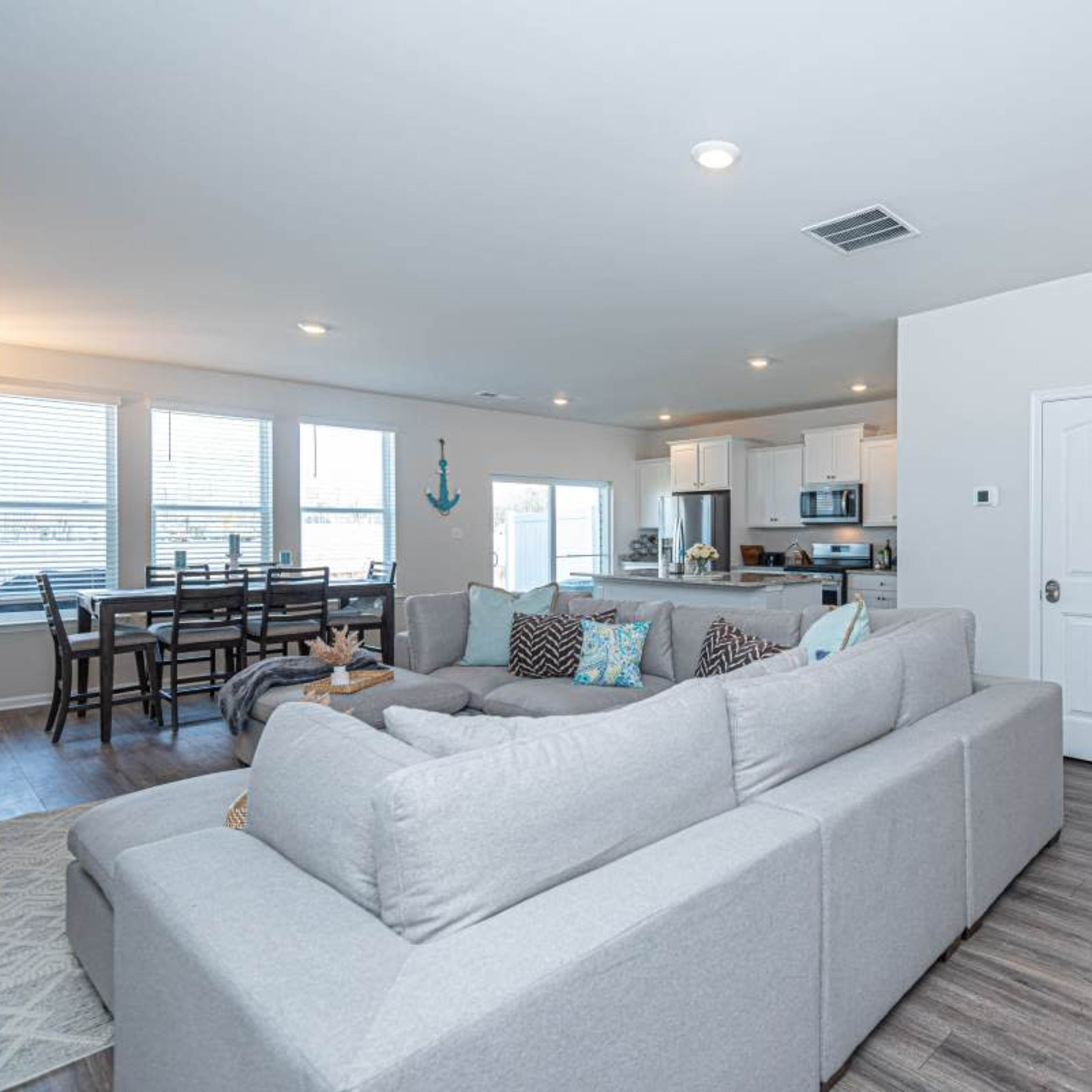A spacious living area features a light-colored sectional sofa, a dining table set for six, and a kitchen visible in the background.