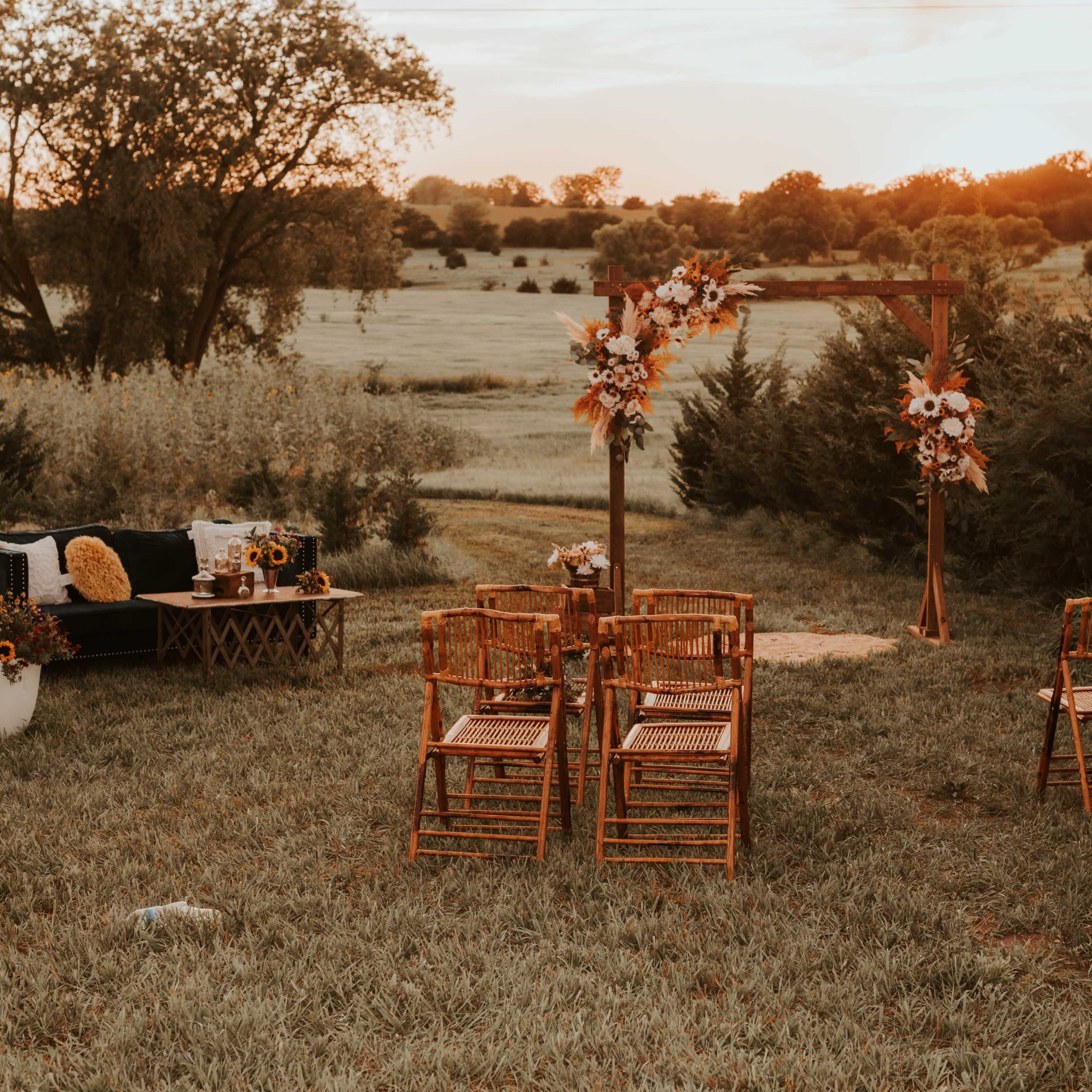 The scene features an outdoor wedding setup with wooden chairs arranged around a decorative arch adorned with flowers, overlooking a grassy field at sunset.