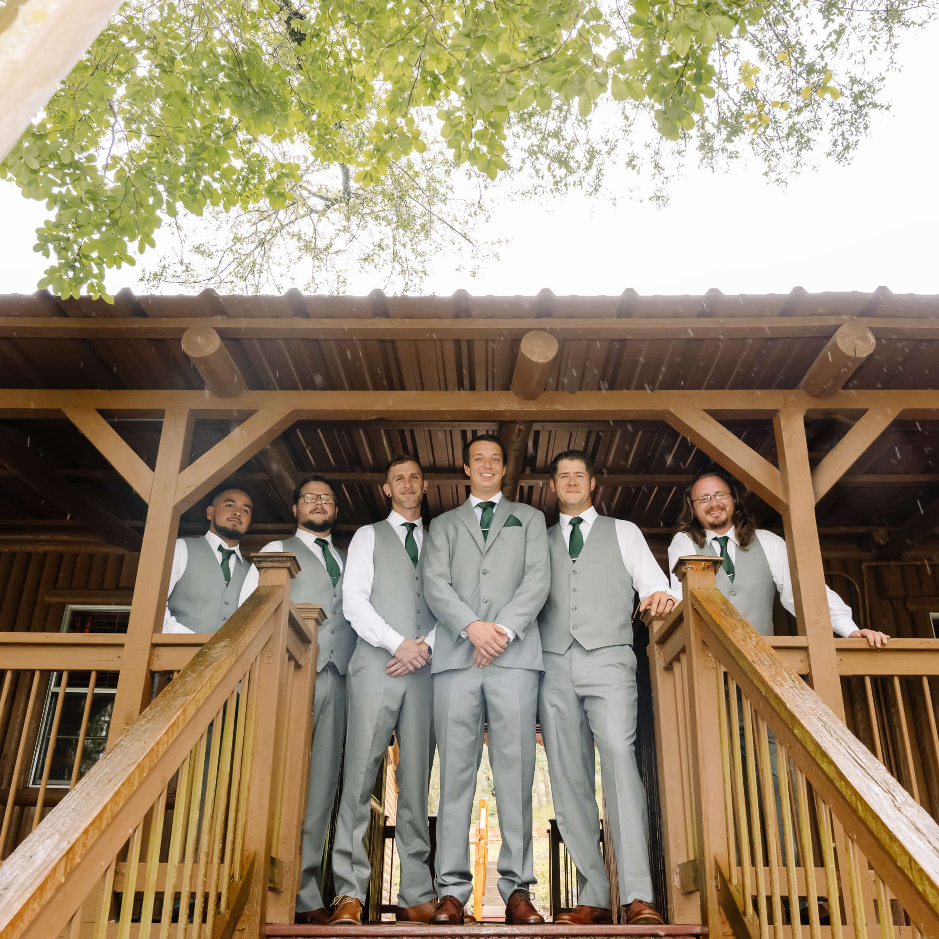 A group of six men in formal attire stands on a wooden staircase in front of a rustic building.