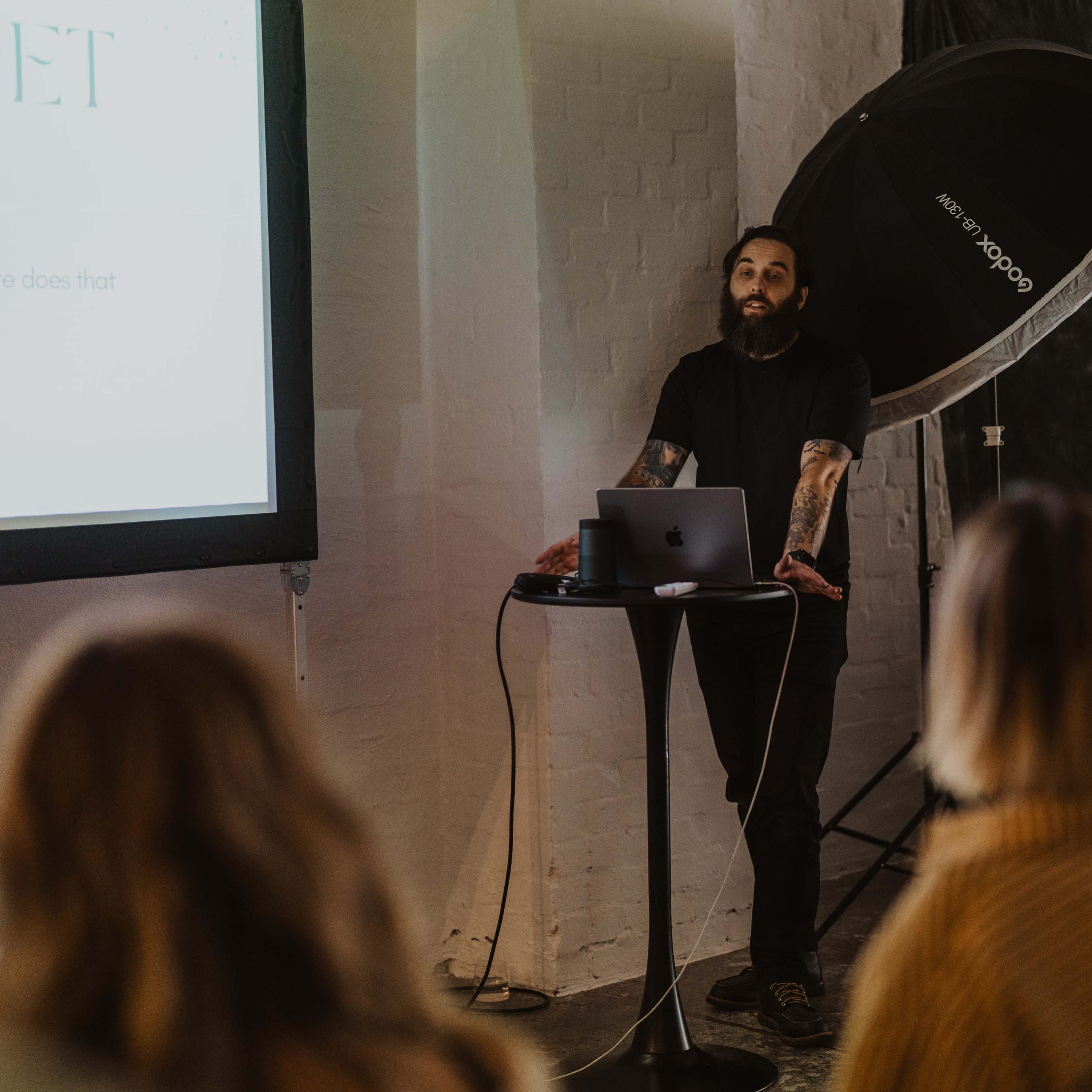 A man with a beard presents in front of an audience, using a laptop and a large screen to display information.