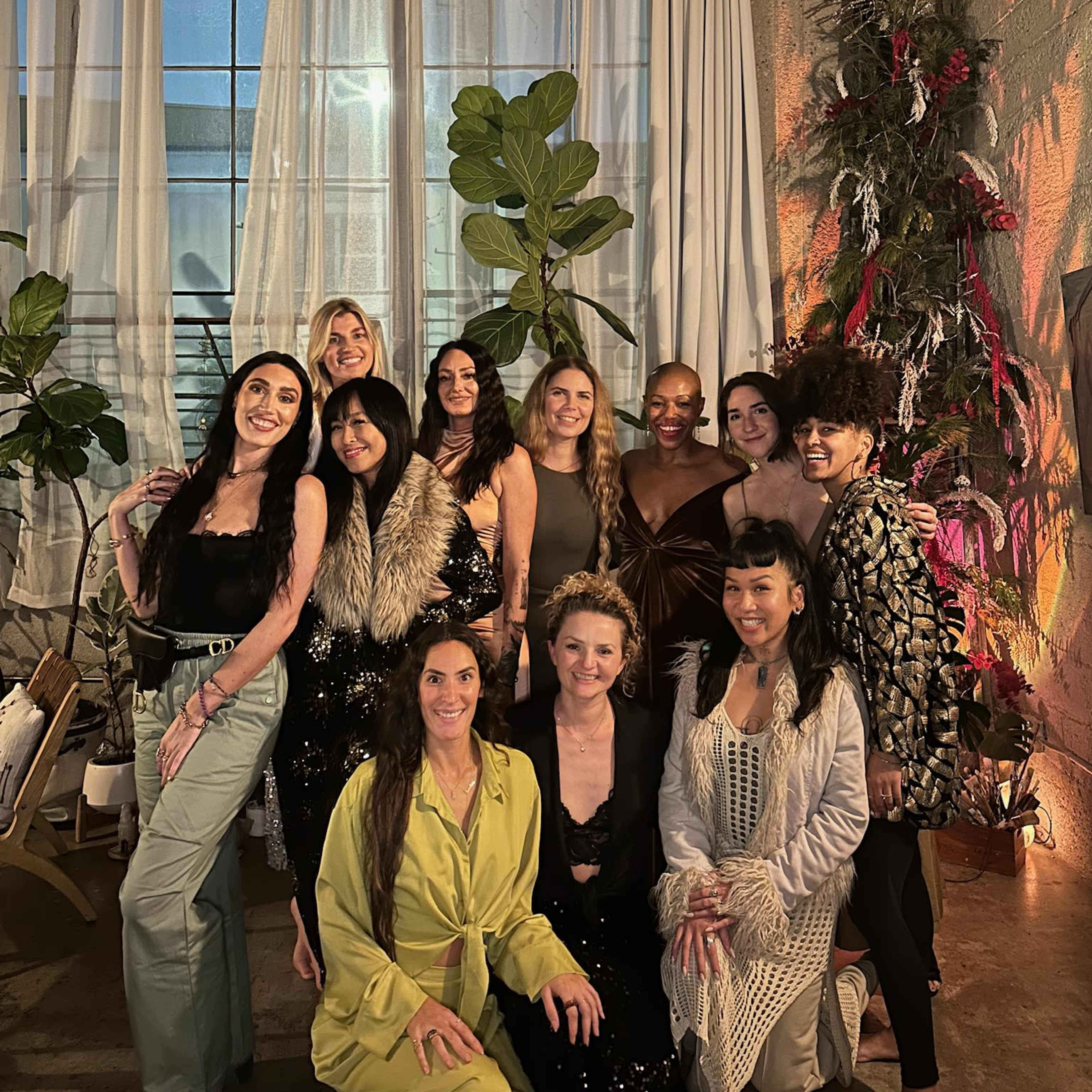 A group of twelve women poses together in a stylish indoor setting adorned with plants and decorative elements.