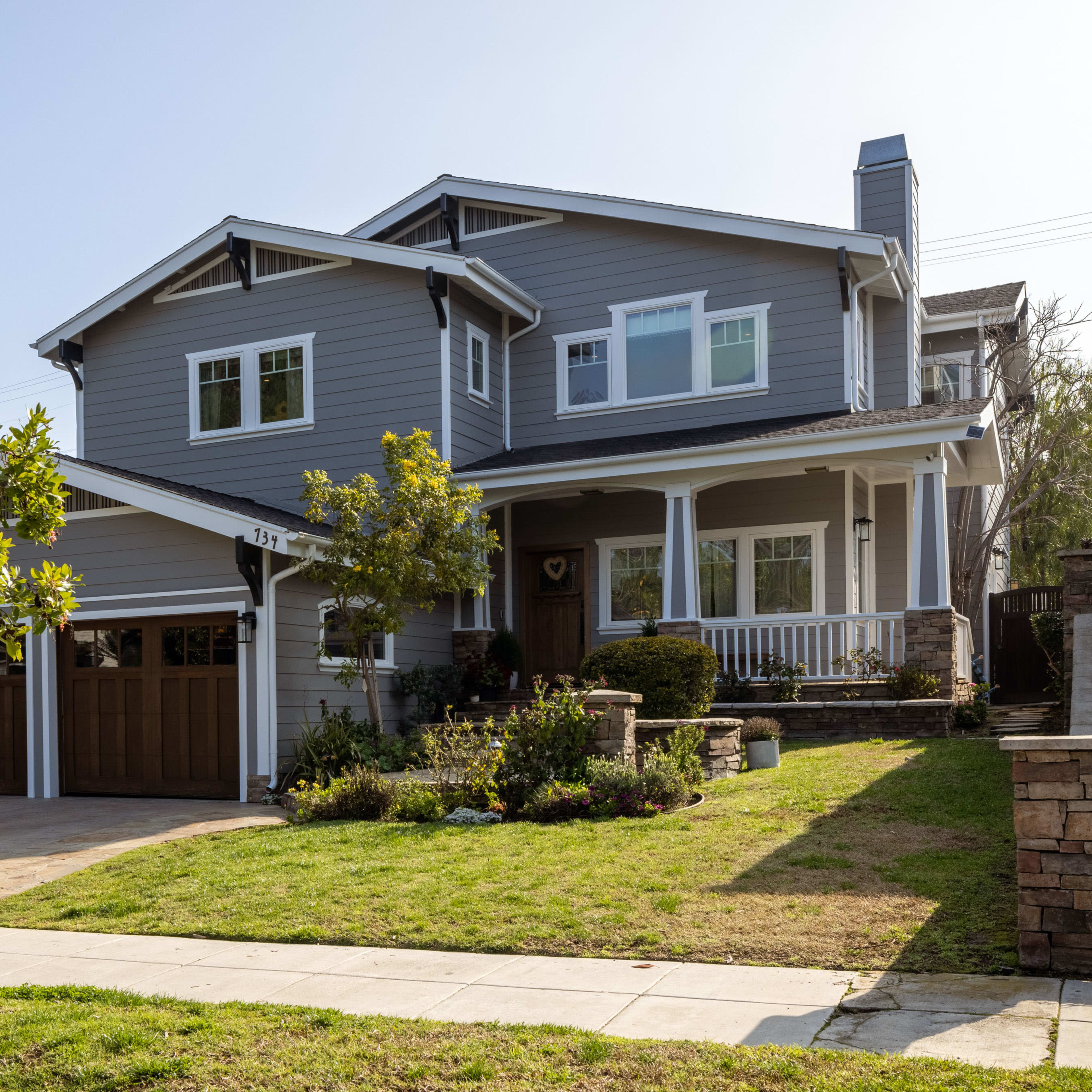 A two-story gray house with a wooden garage door and a front porch is situated on a landscaped yard.