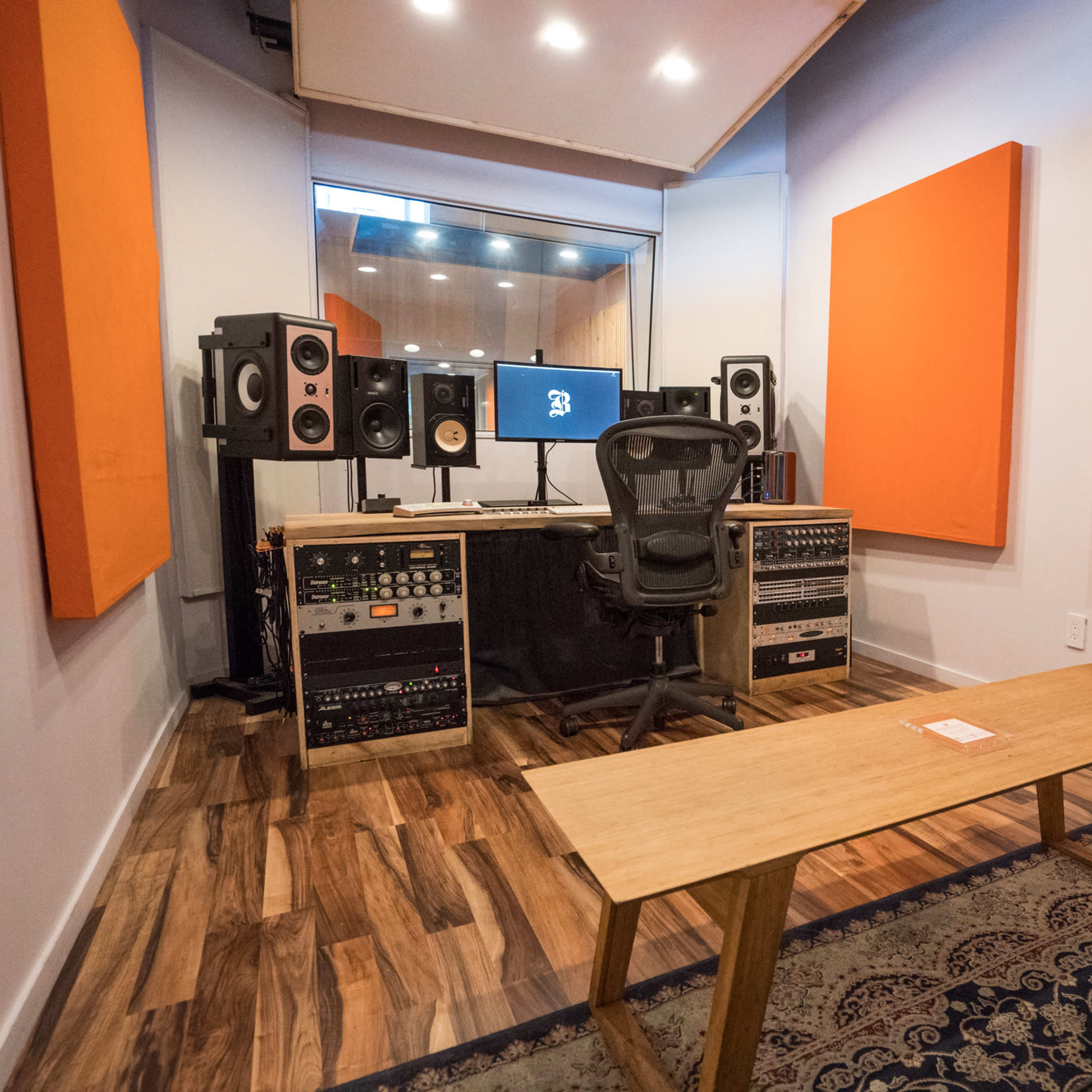 A soundproof recording studio with orange acoustic panels, a mixing desk, studio monitors, and a central chair facing computer screens.