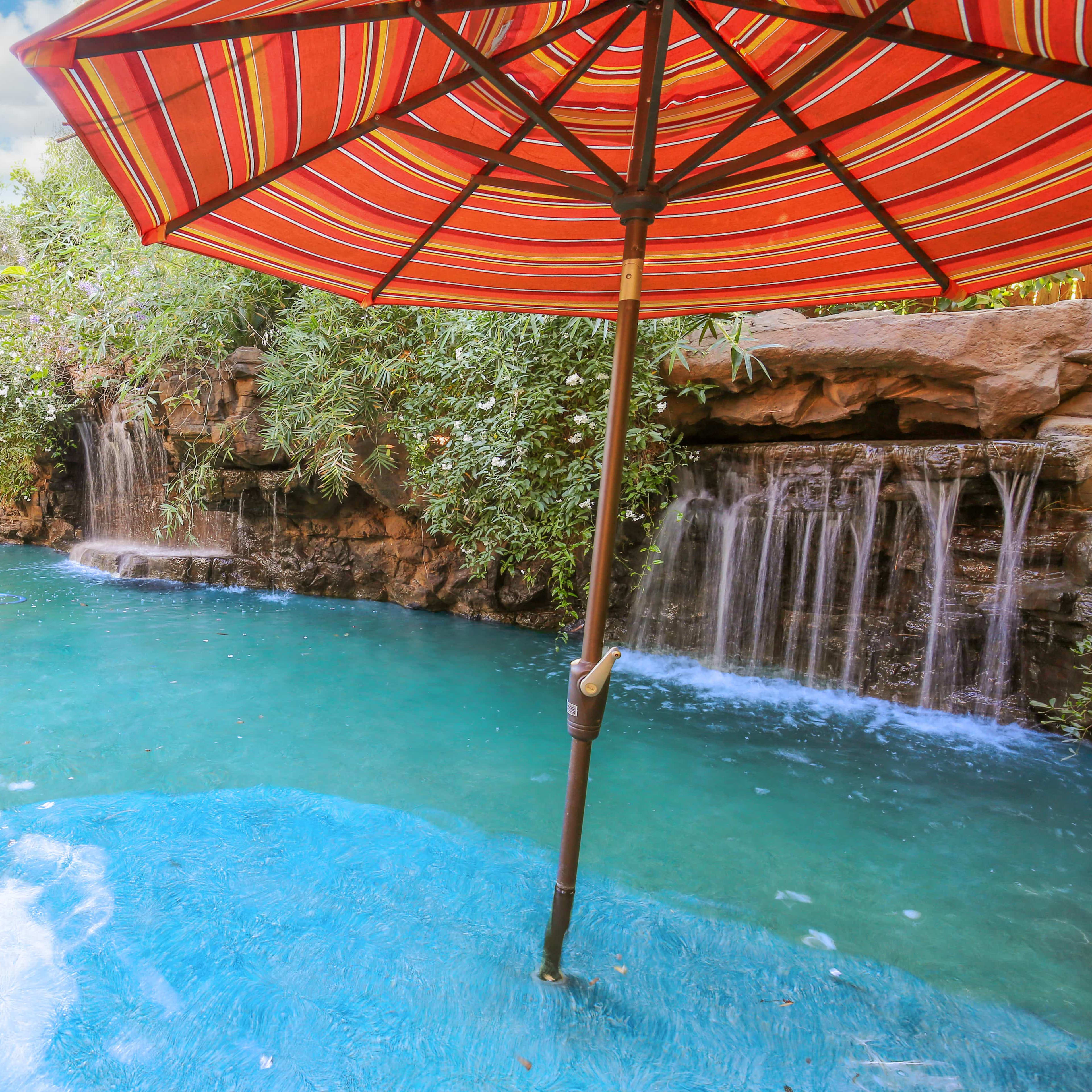 A vibrant pool area with a striped umbrella, surrounded by lush greenery and two small waterfalls cascading into the water.