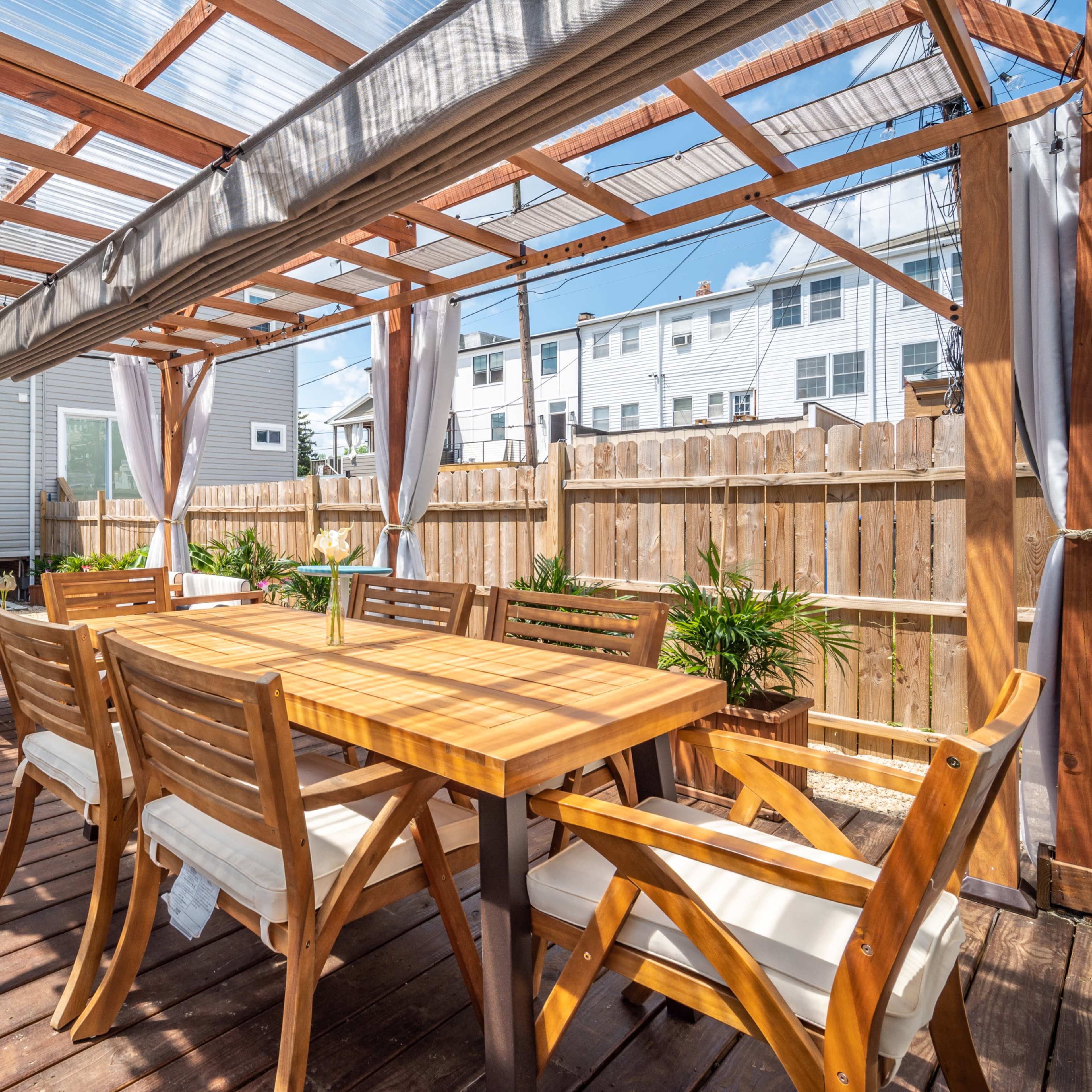 A wooden patio with a dining table and chairs, surrounded by plants and a pergola.