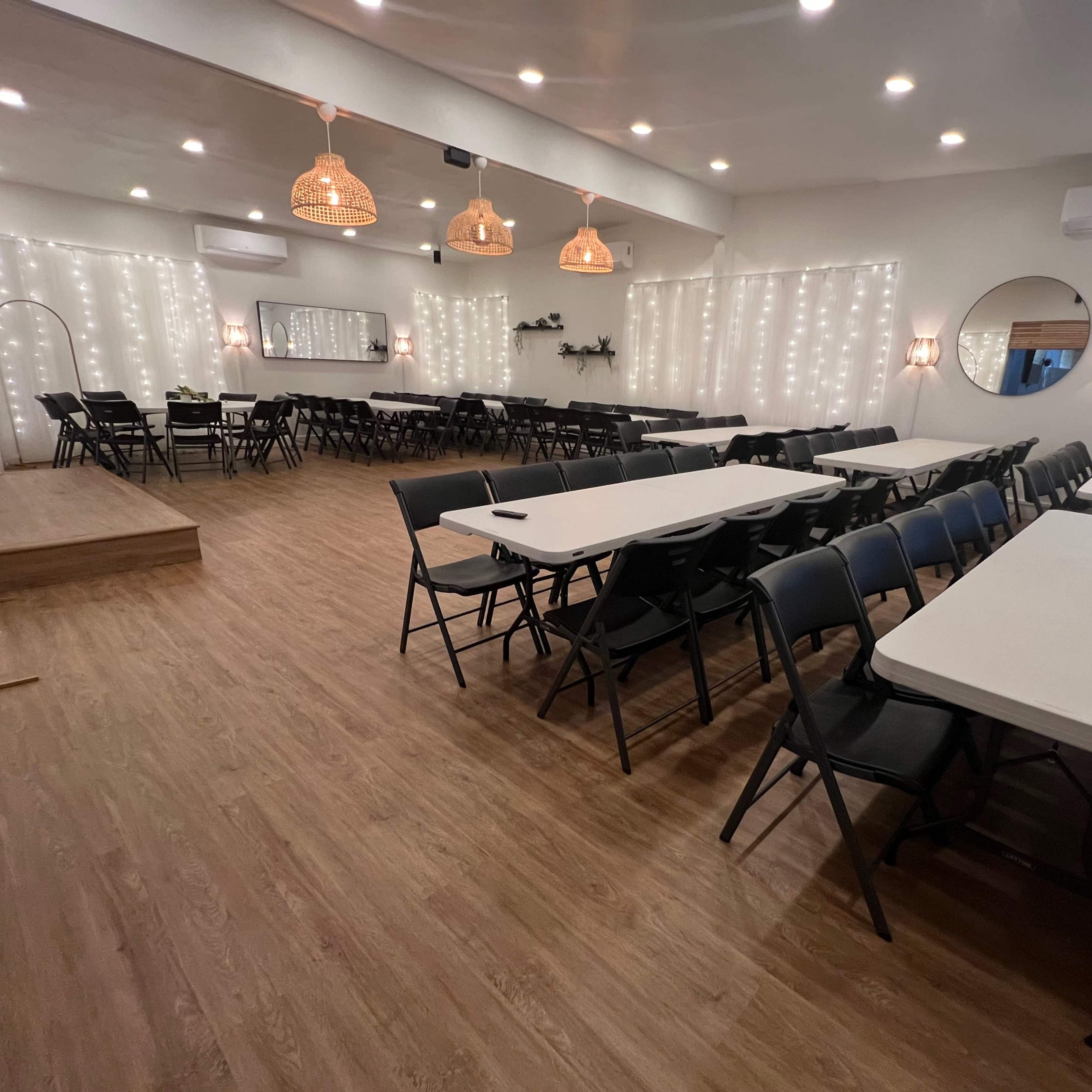 A spacious event room arranged with tables and chairs, featuring string lights along the walls and a small stage area.