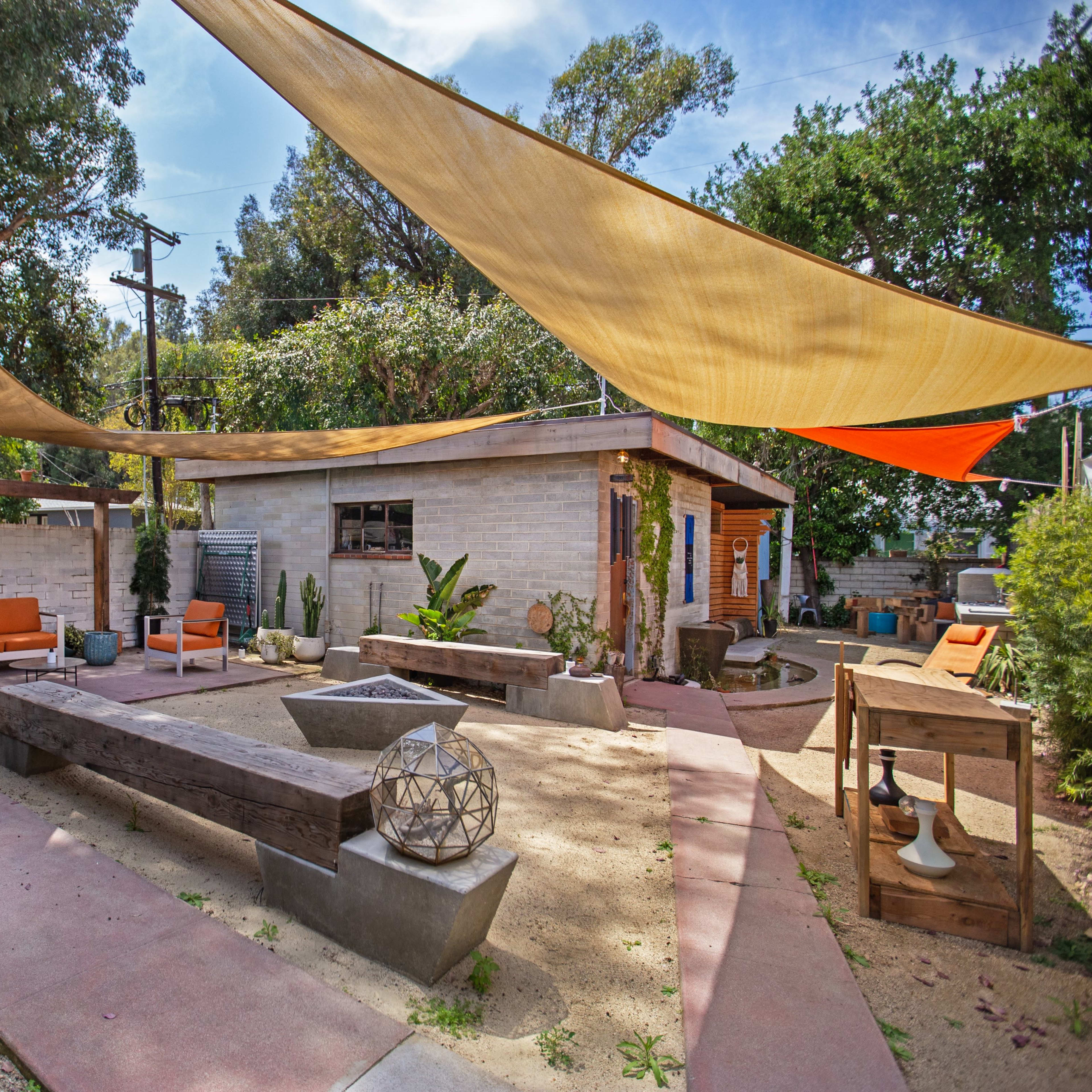 A landscaped outdoor space featuring sandy areas, seating arrangements under large fabric canopies, and a central fire pit surrounded by wooden benches.