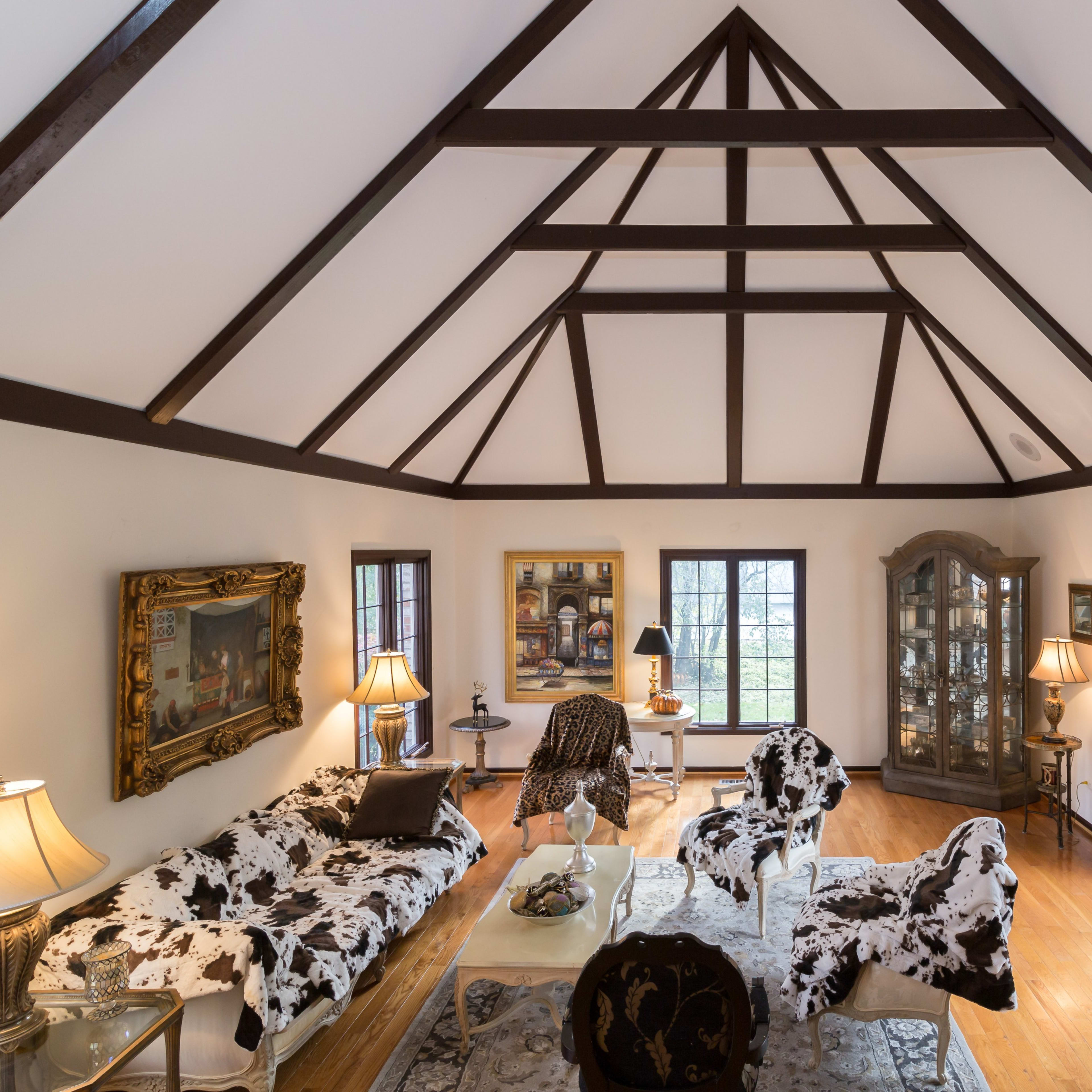 The interior of a spacious living room features a high, exposed beam ceiling, a mix of decorative furniture, and large windows allowing natural light to enter.