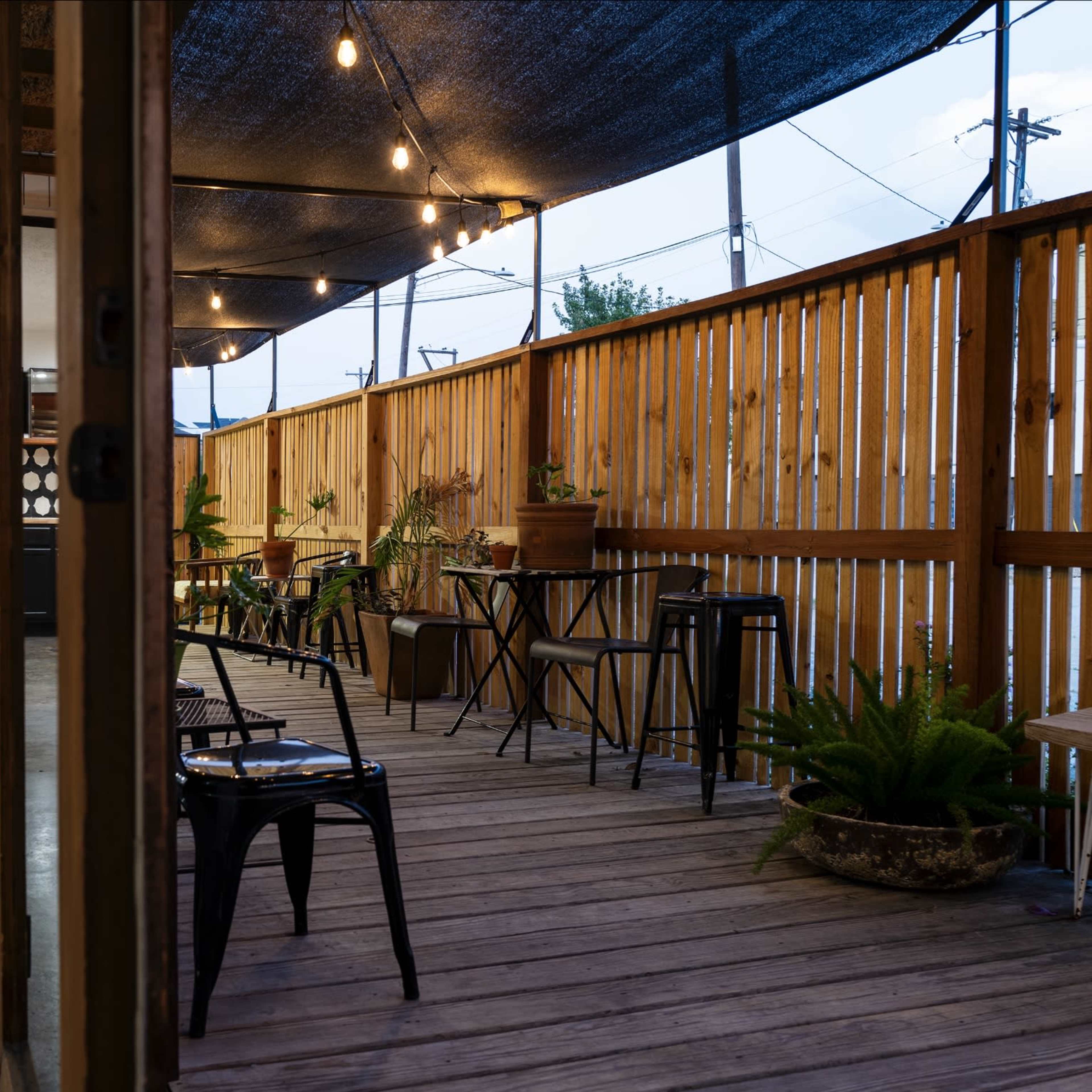 A wooden deck area with string lights and potted plants, leading to an outdoor kitchen space with black cabinetry and a polka dot backsplash.