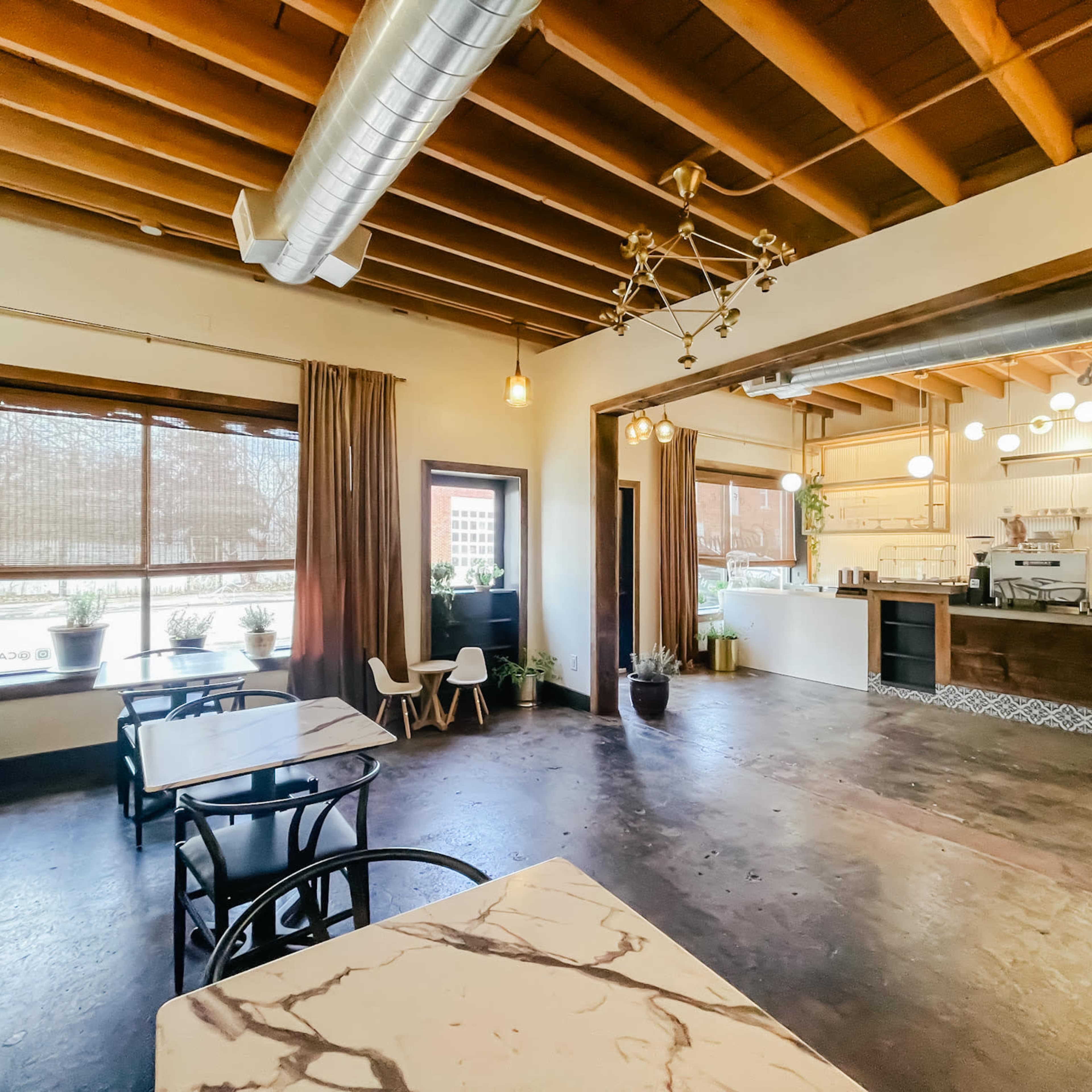 A spacious cafe with wooden beams, marble-topped tables, and large windows allowing natural light to fill the room.