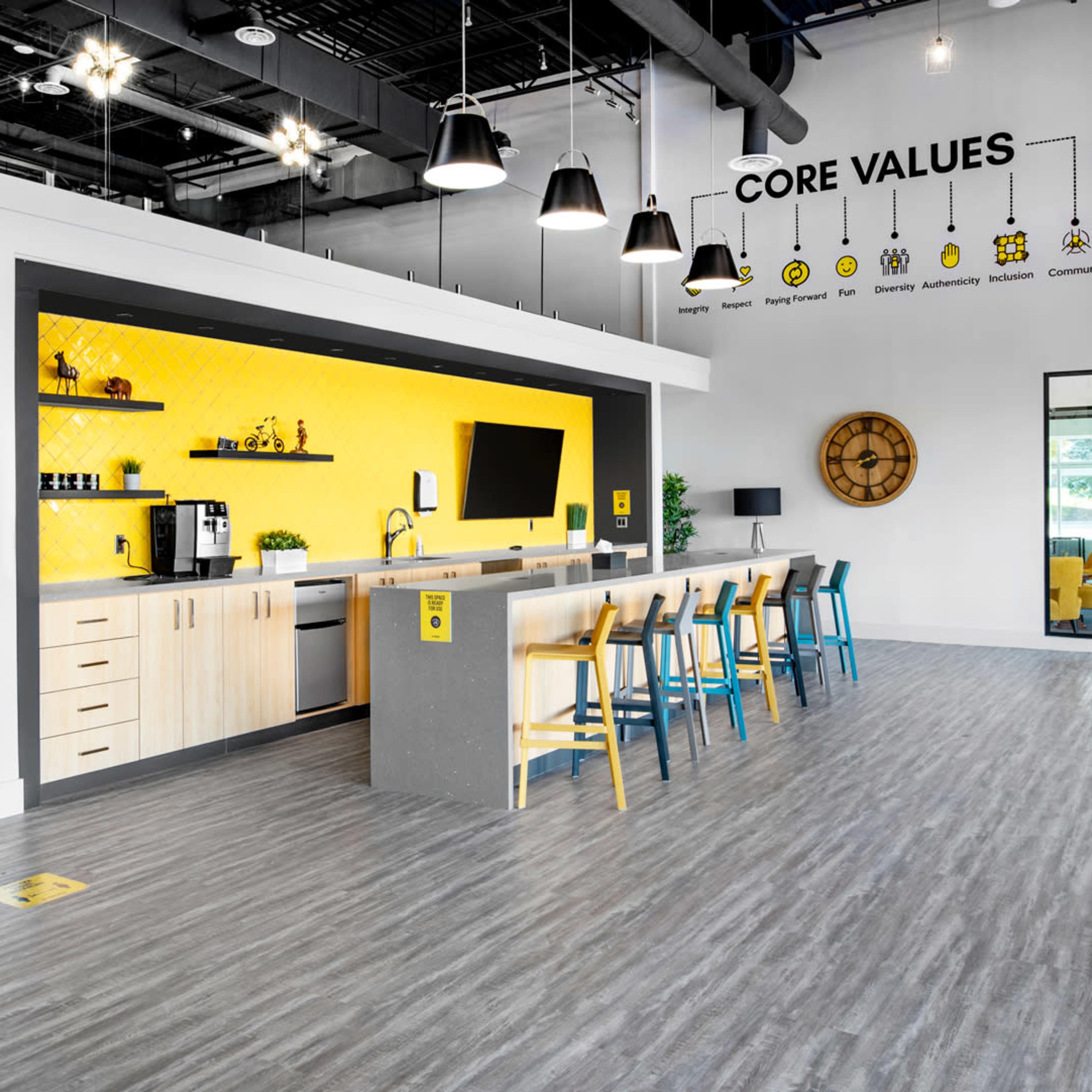 A modern kitchen area with a long bar, bright yellow accent wall, and various appliances, surrounded by colorful stools in a well-lit, open space.