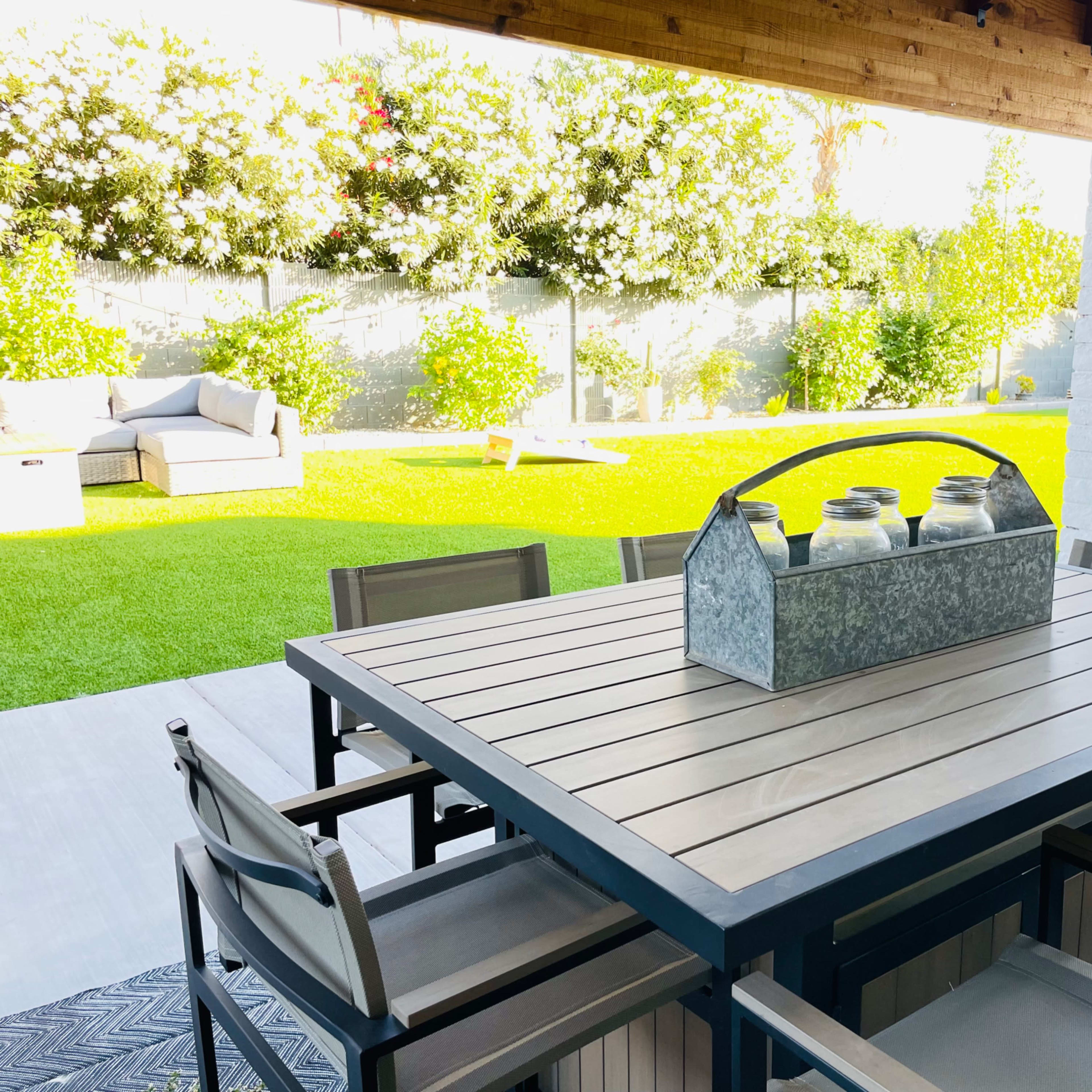 A patio dining table with metal chairs is set under a covered area, overlooking a well-maintained green lawn and shrubbery.