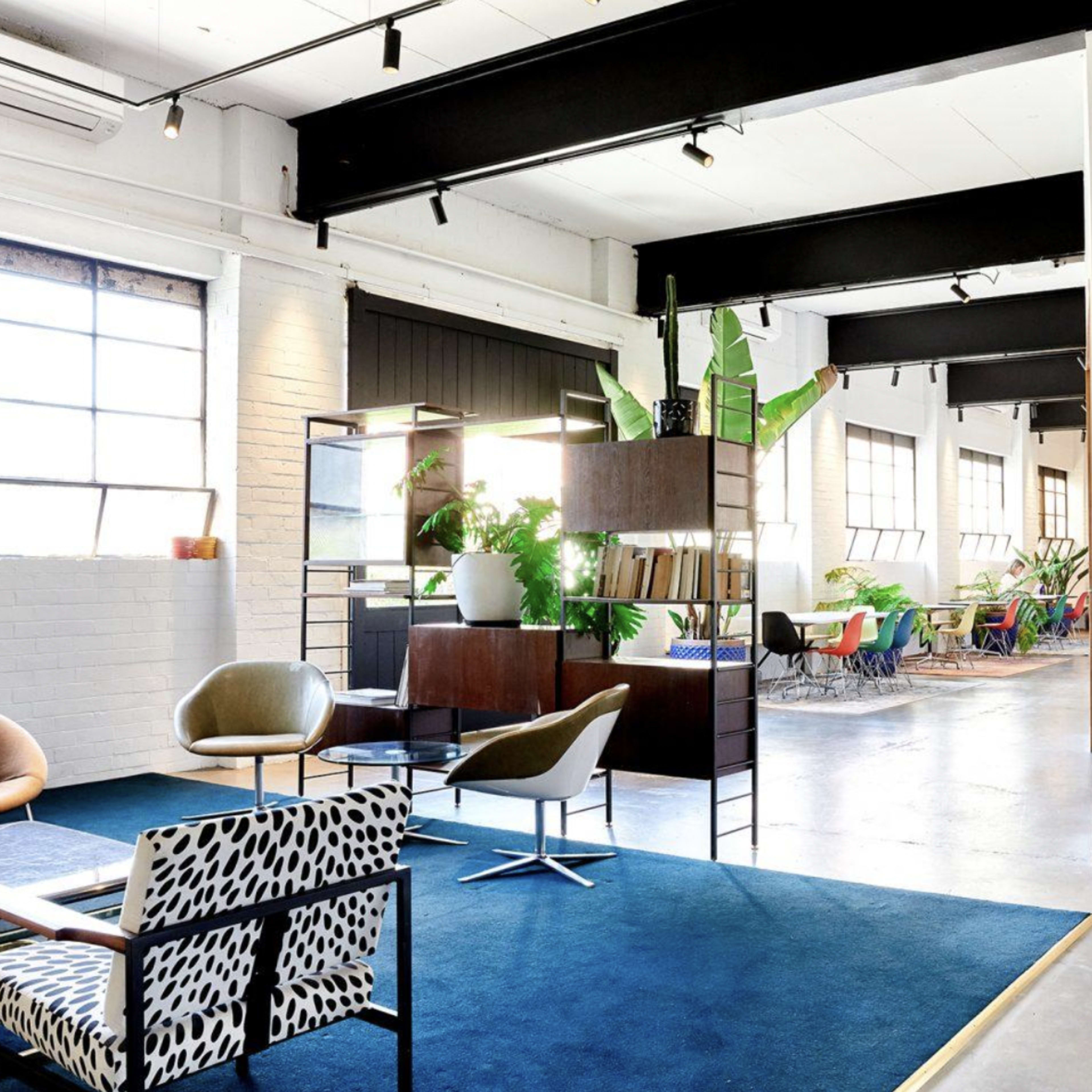 A modern office interior with a combination of seating areas, decorative plants, and large windows allowing natural light to enter.