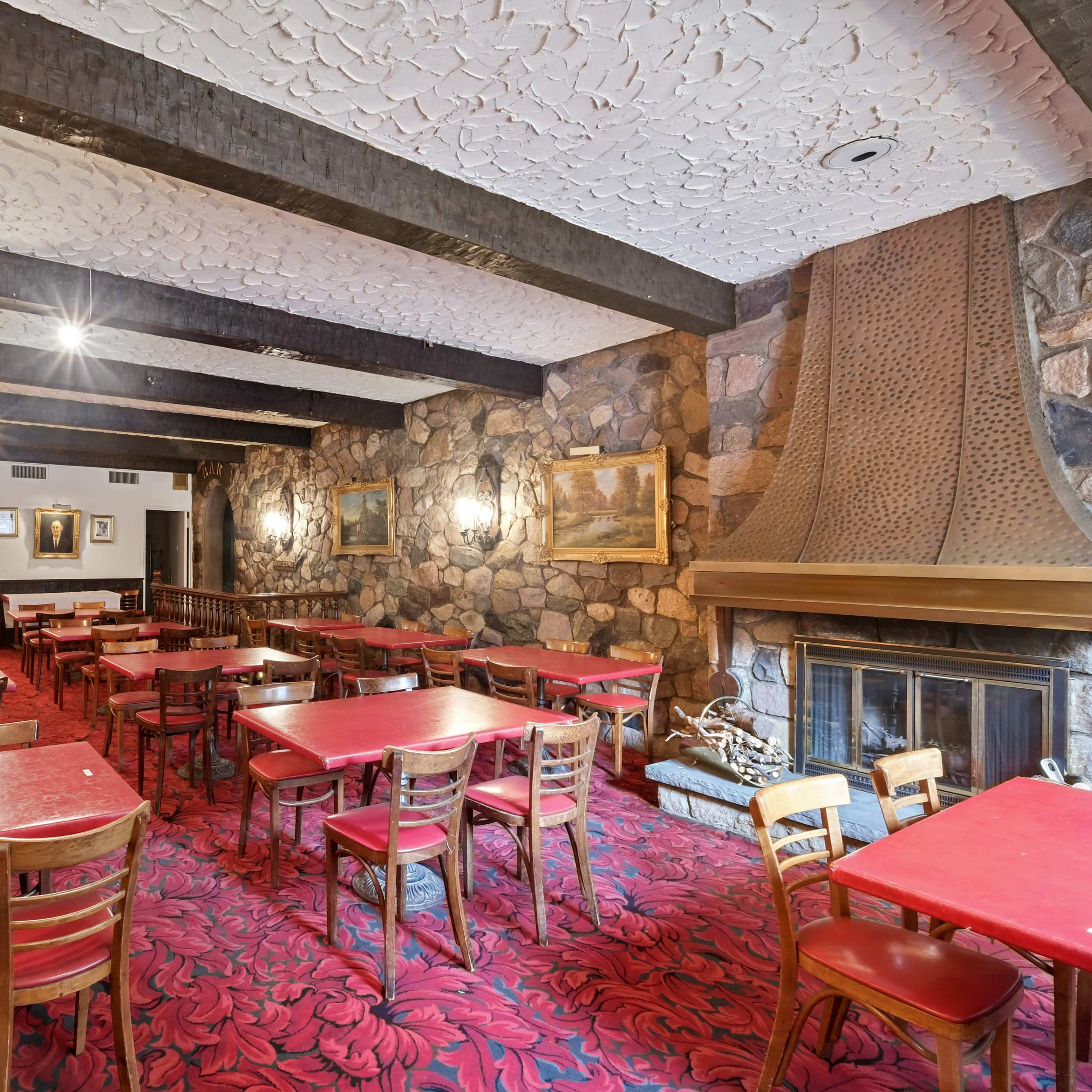 A restaurant interior featuring red tables and chairs, a stone fireplace, and textured walls with wooden beams overhead.