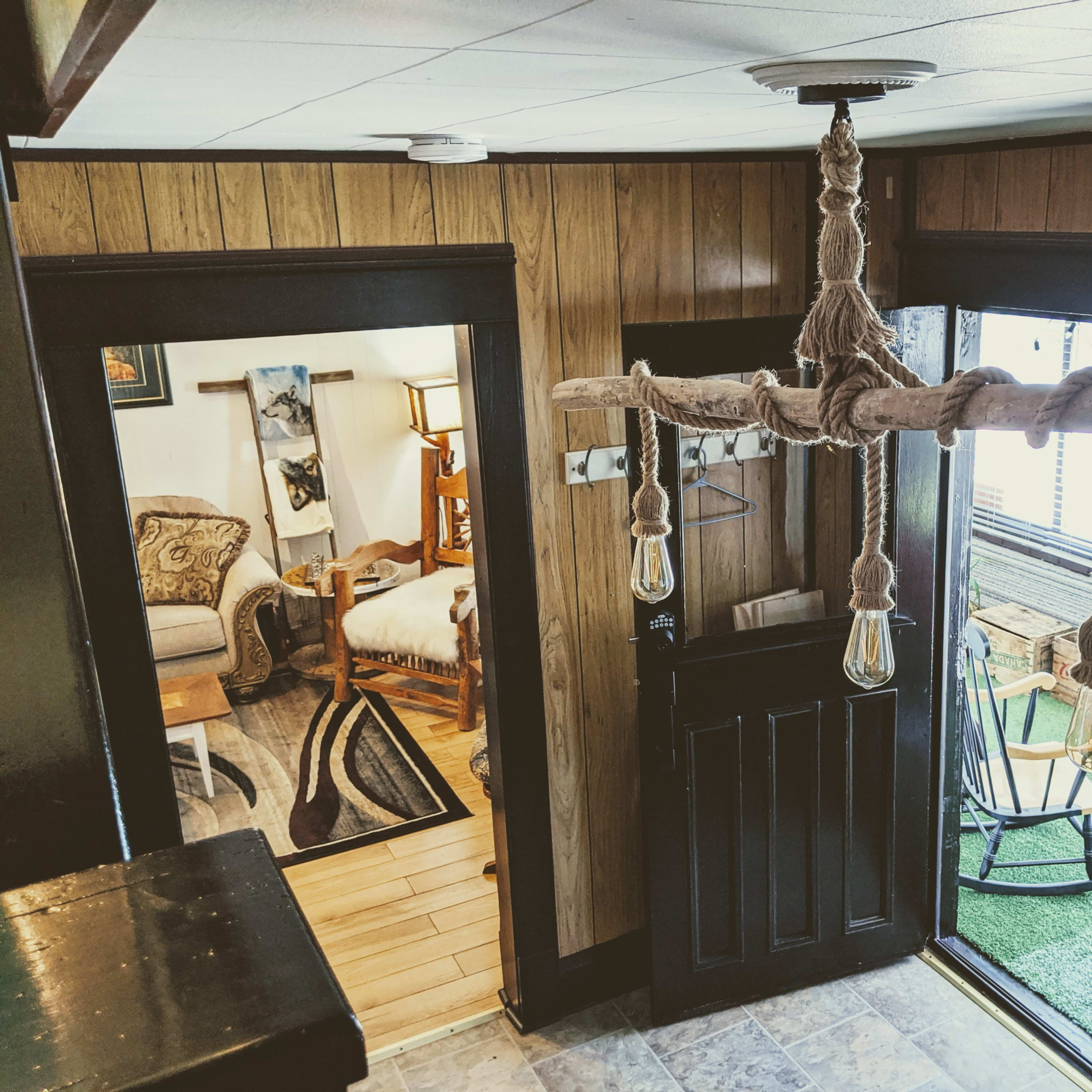 A hallway with a wooden chandelier hanging from the ceiling, leading to two doorways: one into a cozy room with a bed and armchair, and another opening to a small outdoor area with a rocking chair.