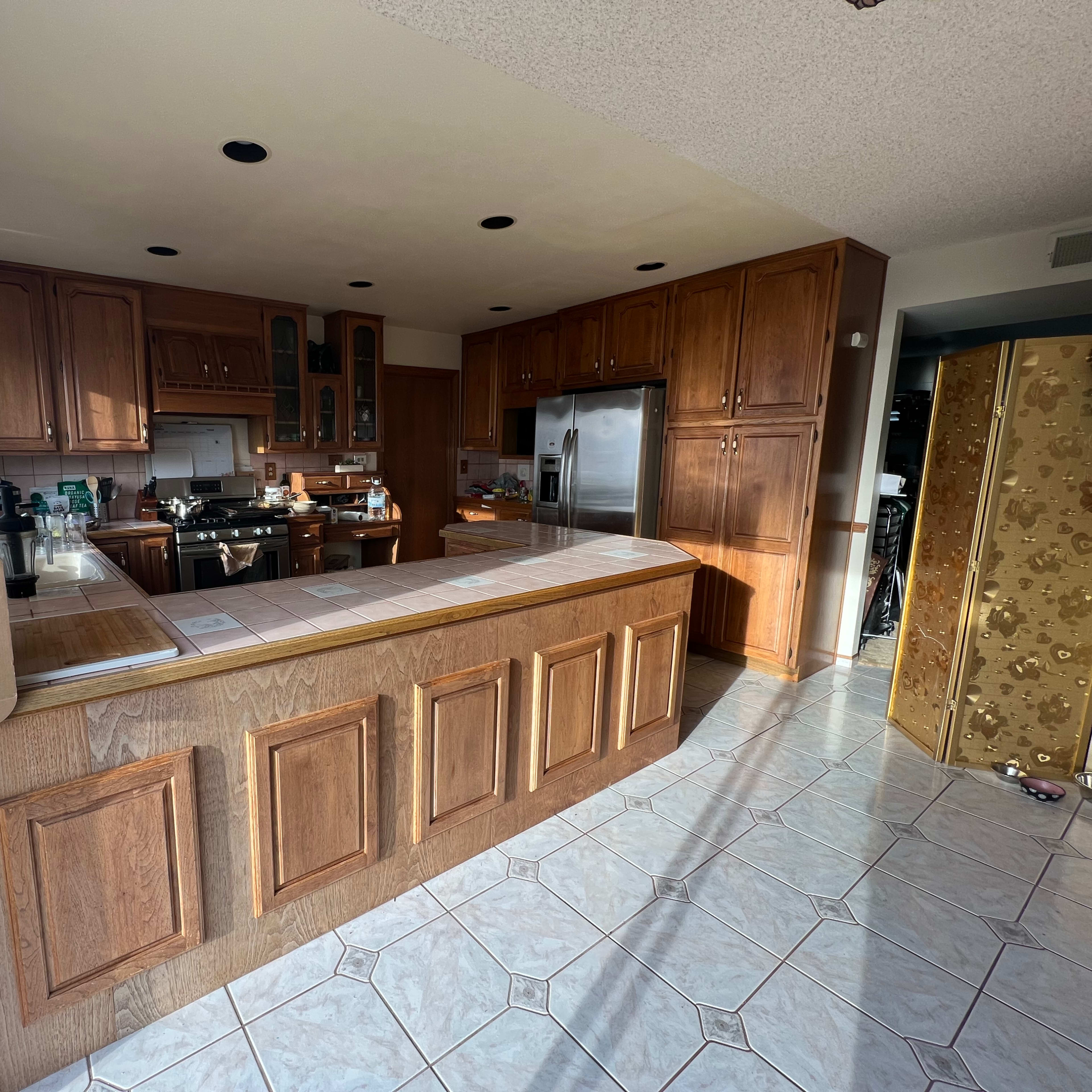 A spacious kitchen with wooden cabinetry, a central island, and tile flooring, illuminated by natural light from large windows.