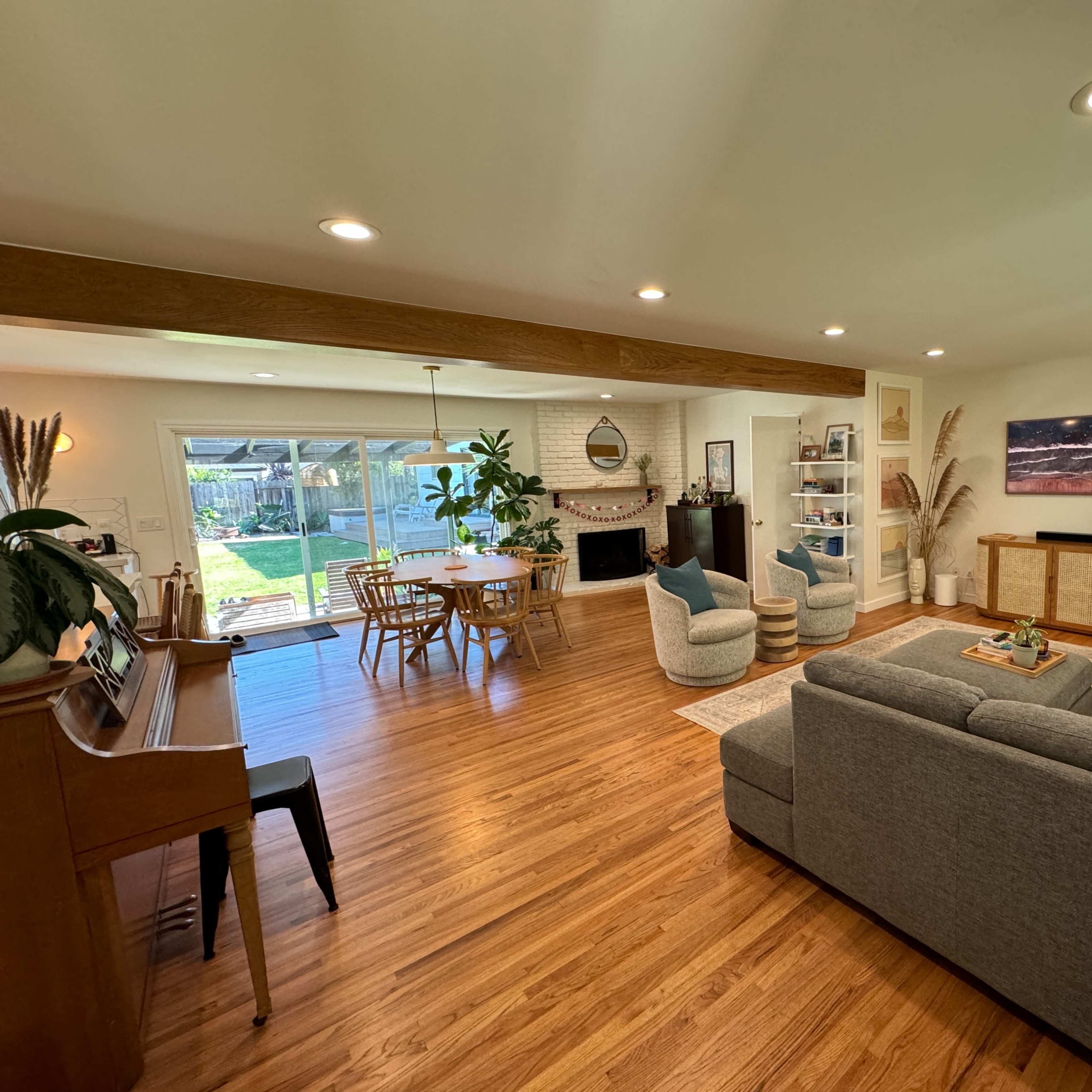 A modern living room with a sectional sofa, piano, and a dining area that opens to a backyard through large glass doors.