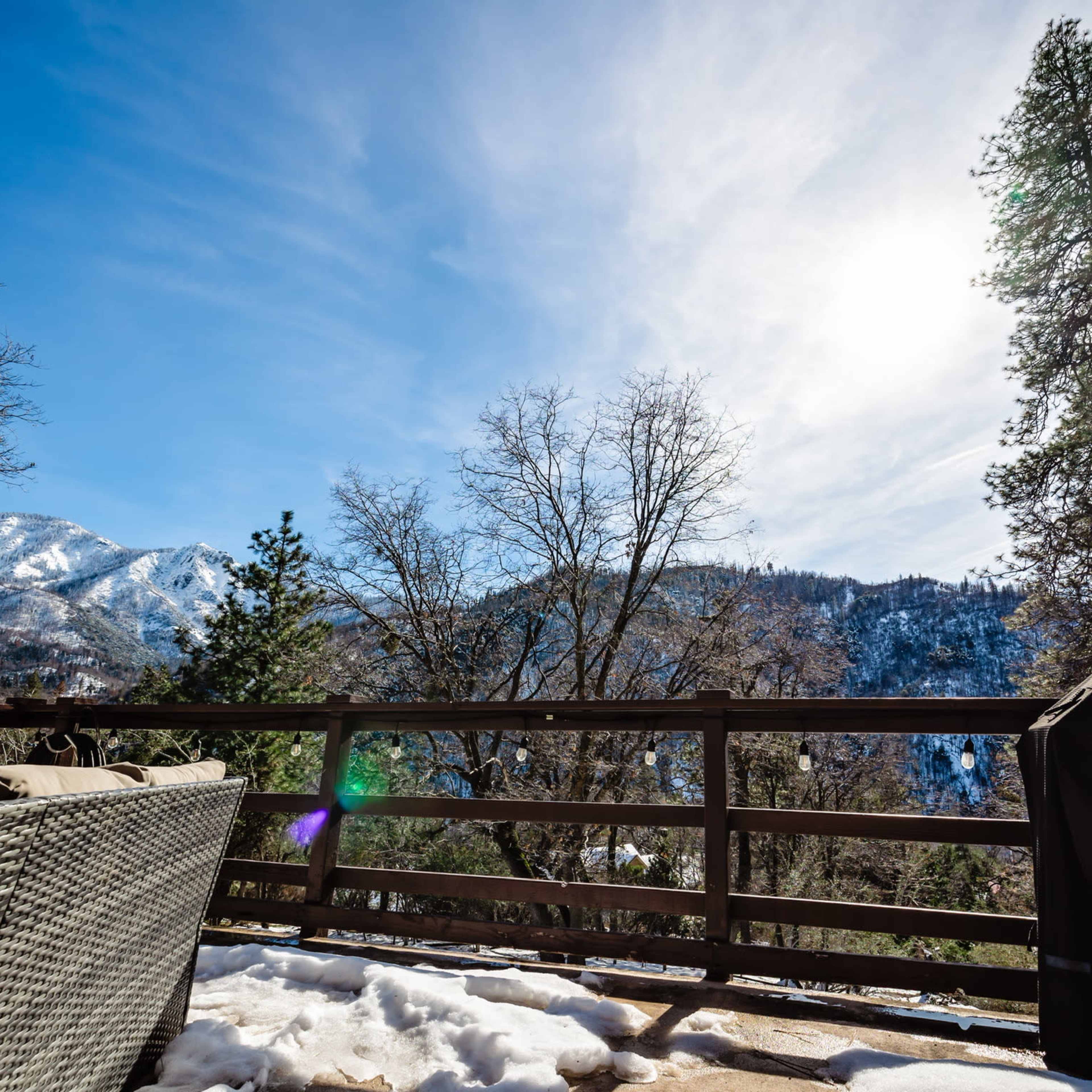 A view from a wooden deck featuring a barbecue grill and surrounded by snow-covered ground, with mountains and trees in the background under a clear blue sky.
