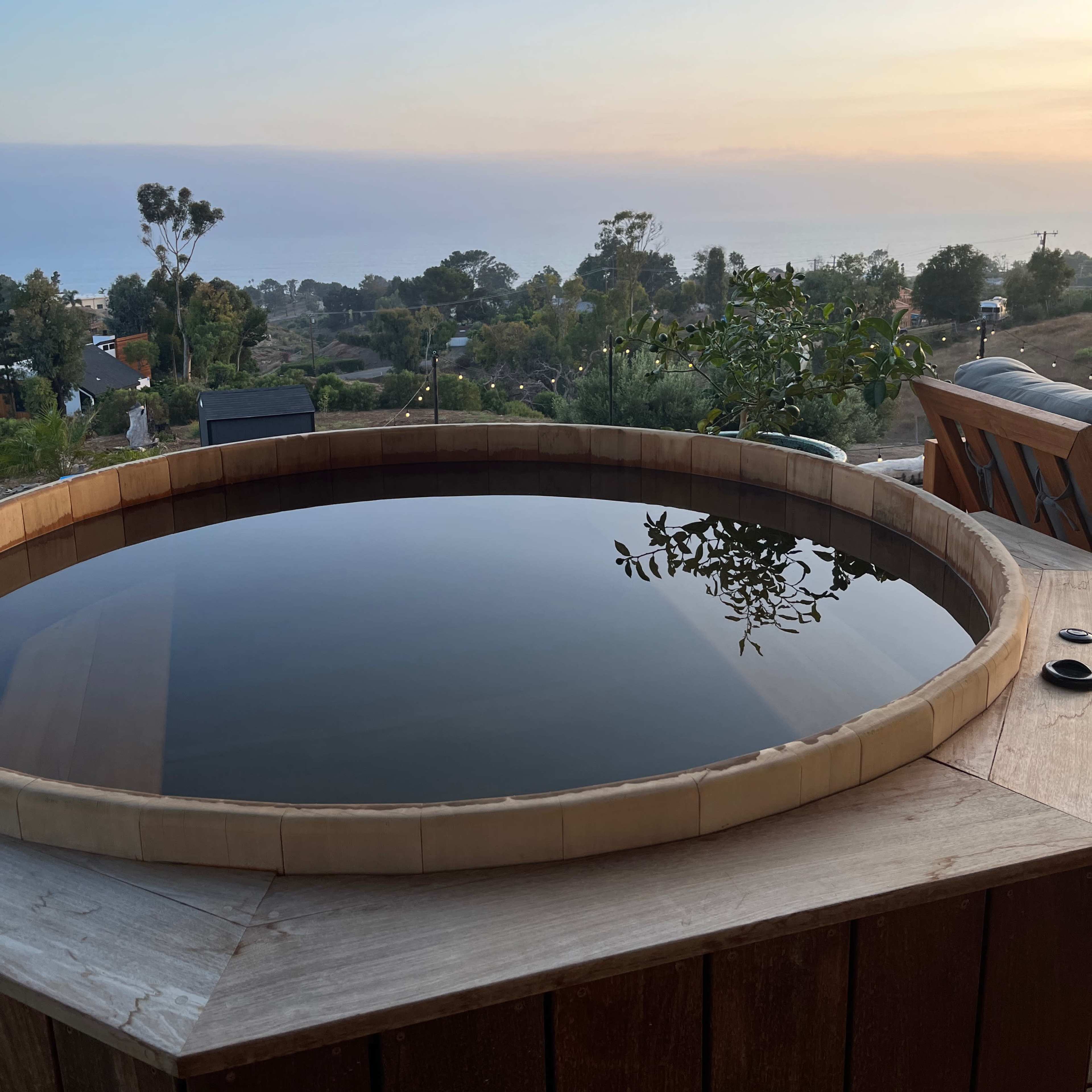 A wooden hot tub surrounded by a scenic view of hills, trees, and a sunset sky.