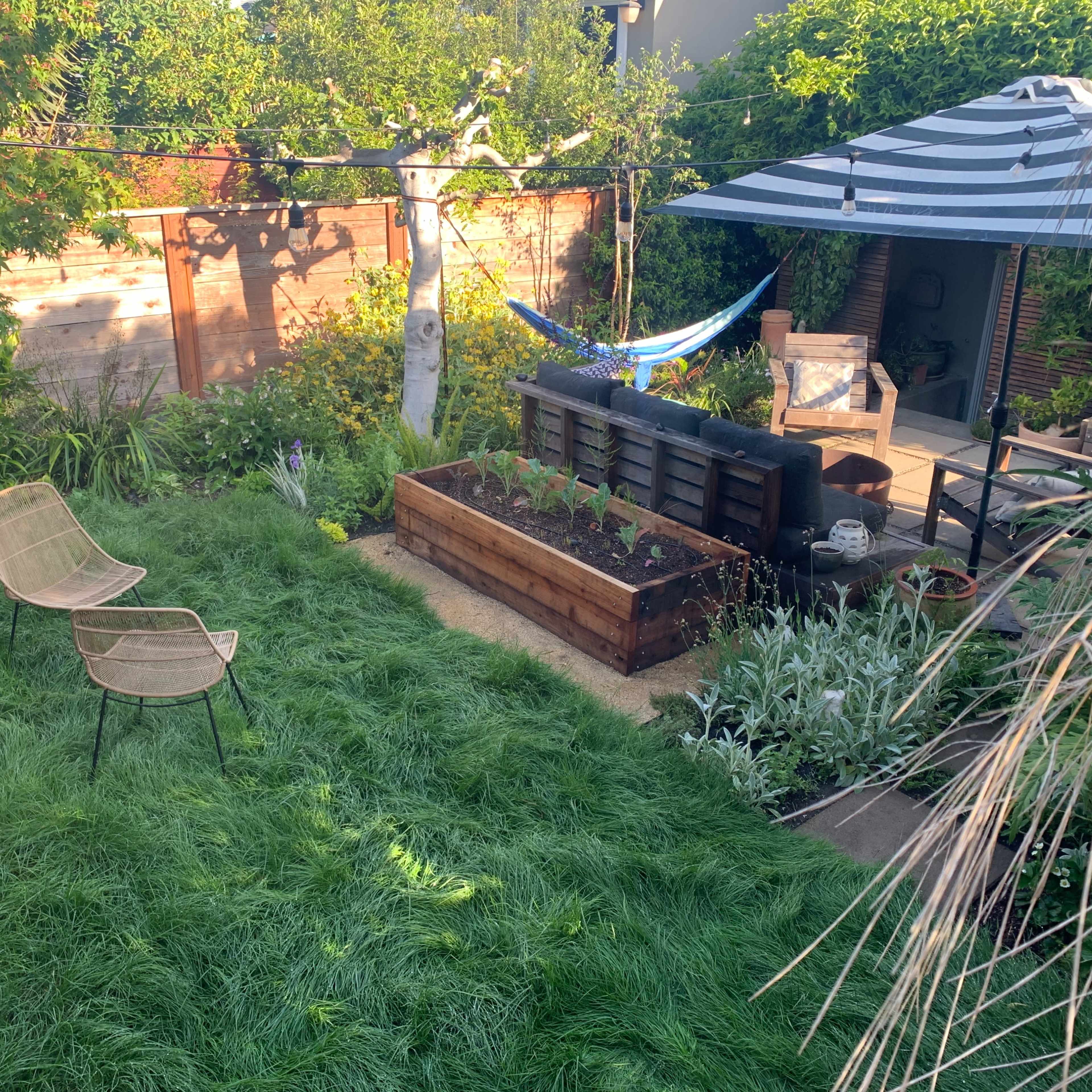 A backyard garden features a striped umbrella over a seating area, a hammock, and a raised garden bed surrounded by lush greenery.