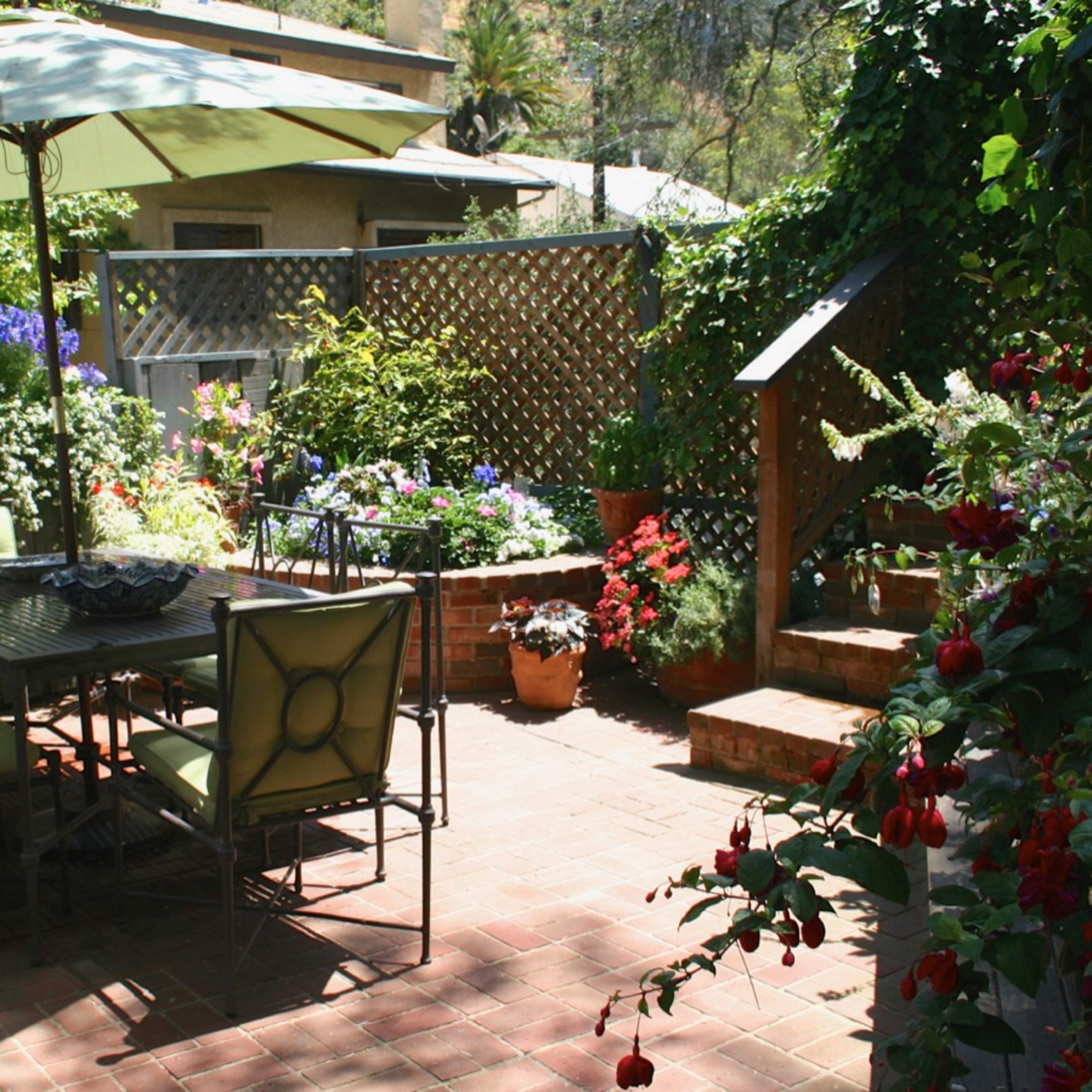 A patio with a dining table and chairs is surrounded by colorful flower pots and plants, with an umbrella providing shade.