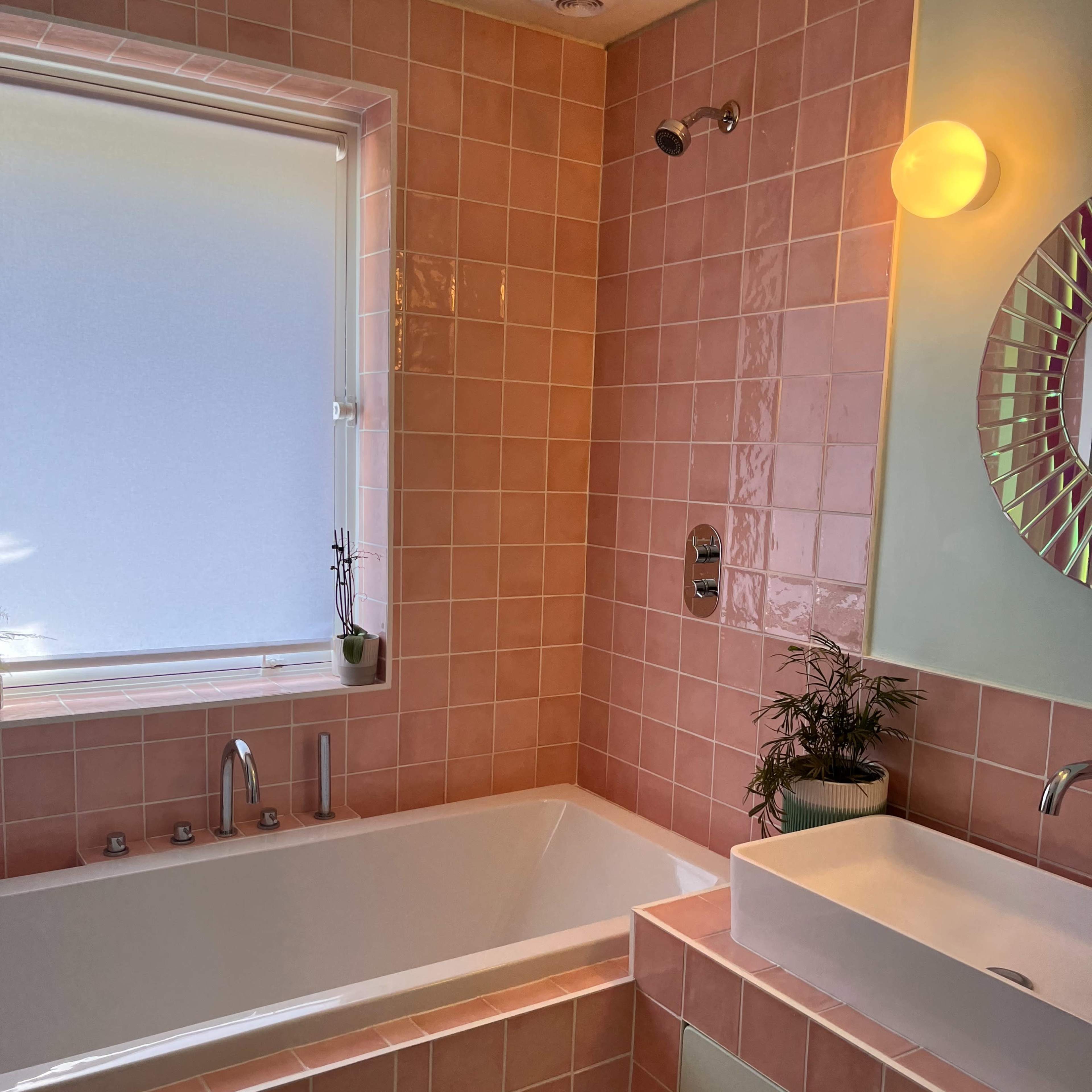 A bathroom with pink tiled walls, a white bathtub, a modern sink, and a window letting in natural light.